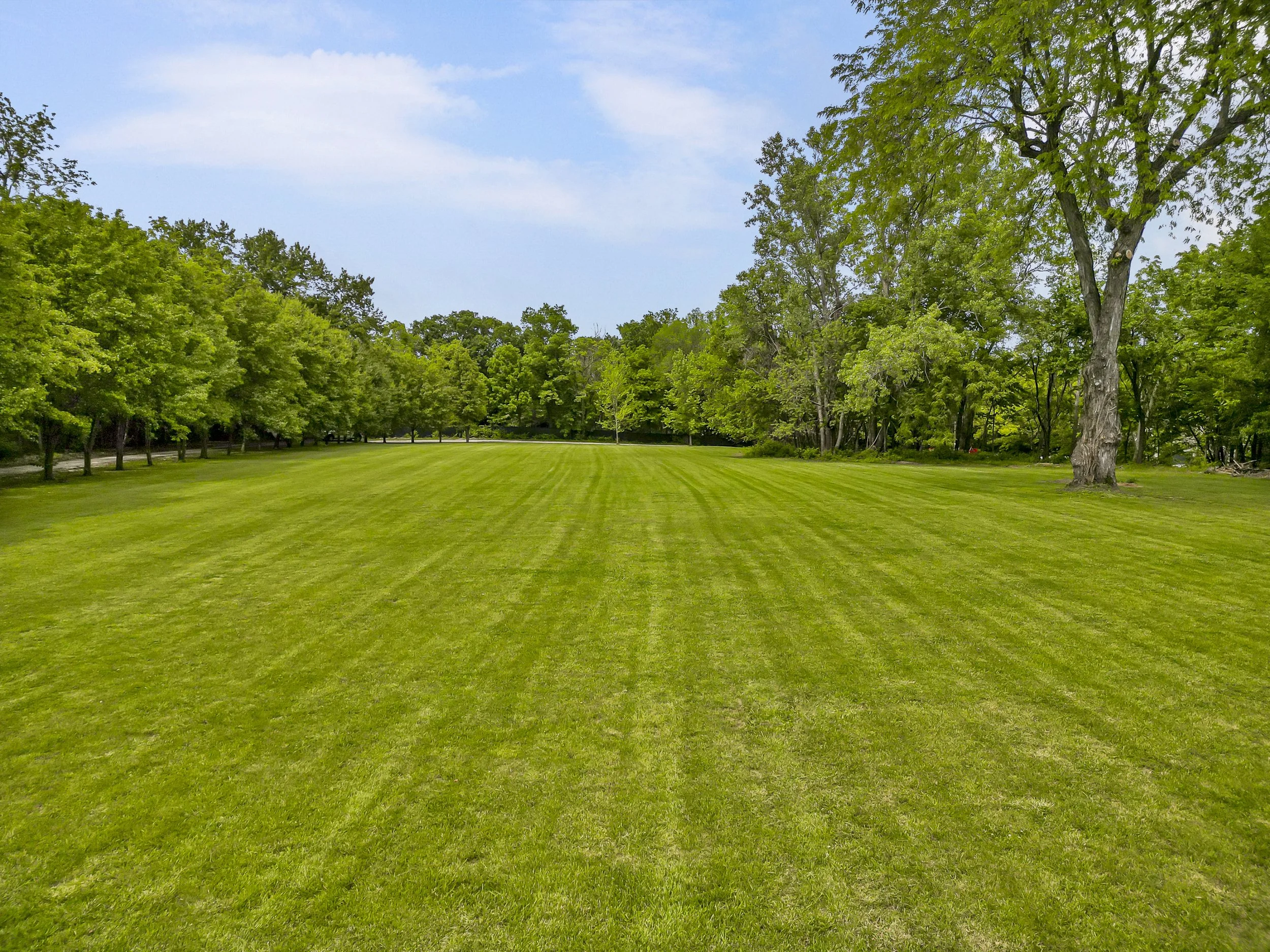 A large grassy field with lines of mowed grass, surrounded by green trees and a partly cloudy sky.