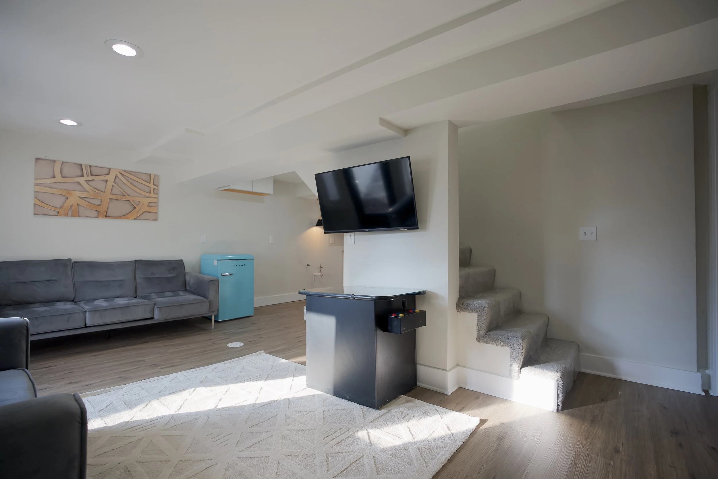 Living room with gray couches, TV mounted on the wall, cream stairs, wooden floor, and white walls.