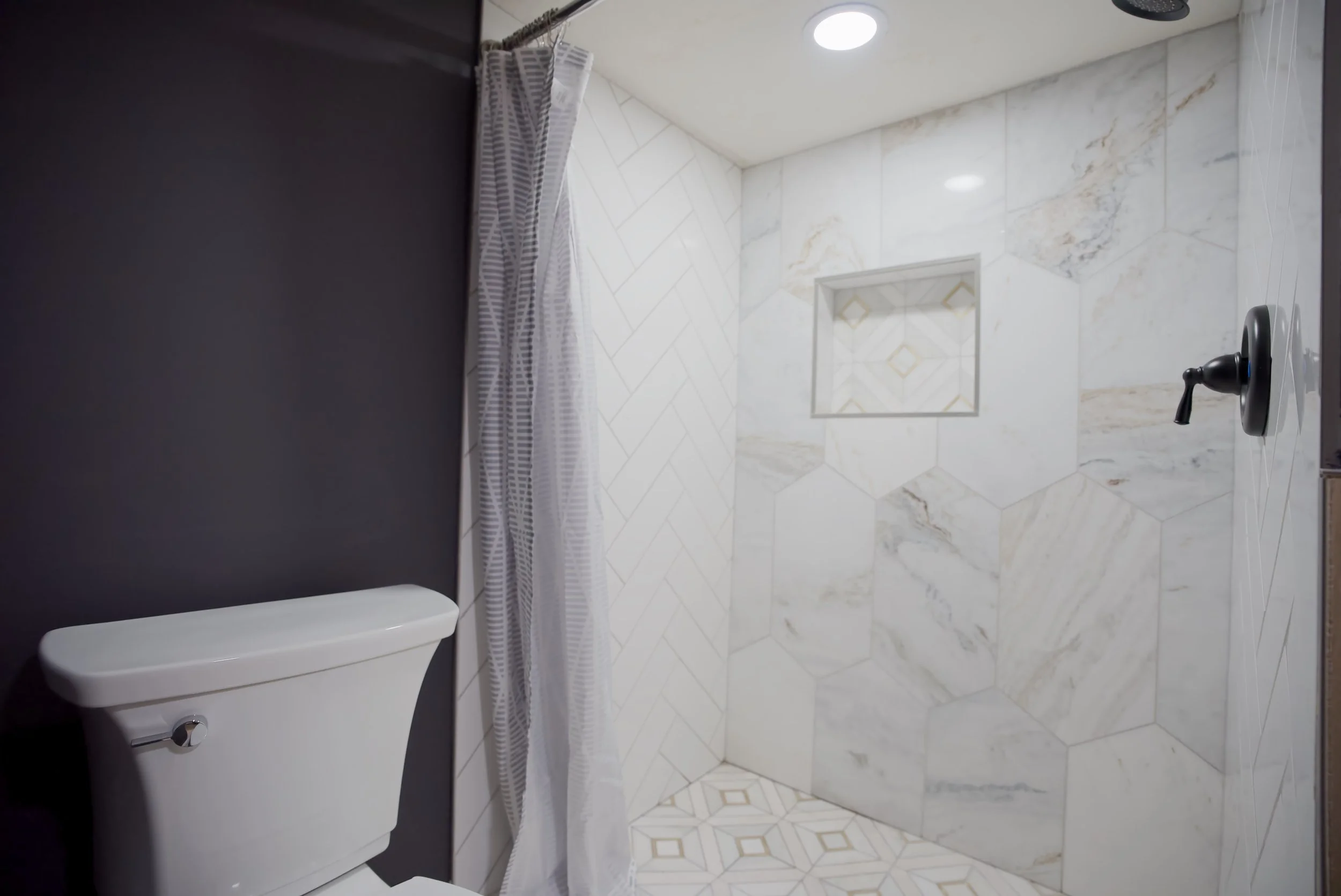 Bathroom shower with white marble tiles, built-in shelf, black showerhead, shower curtain, toilet to the left, and a black wall to the left side.