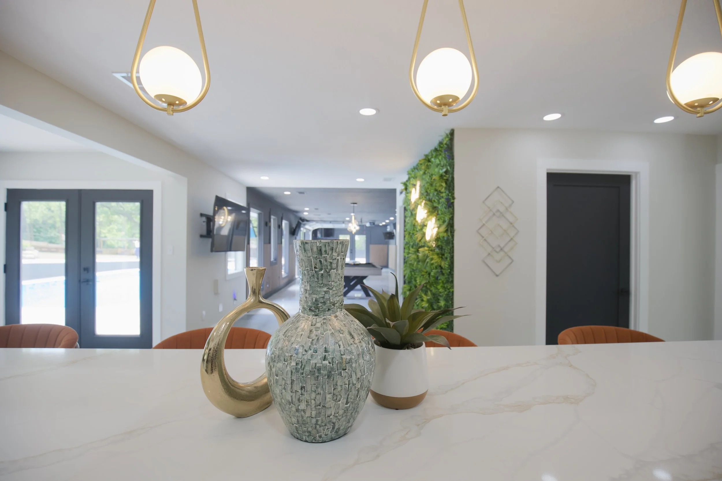 Decorative vases and a plant on a white countertop in a modern living space with hanging lights, a living area with chairs, and a green wall in the background.