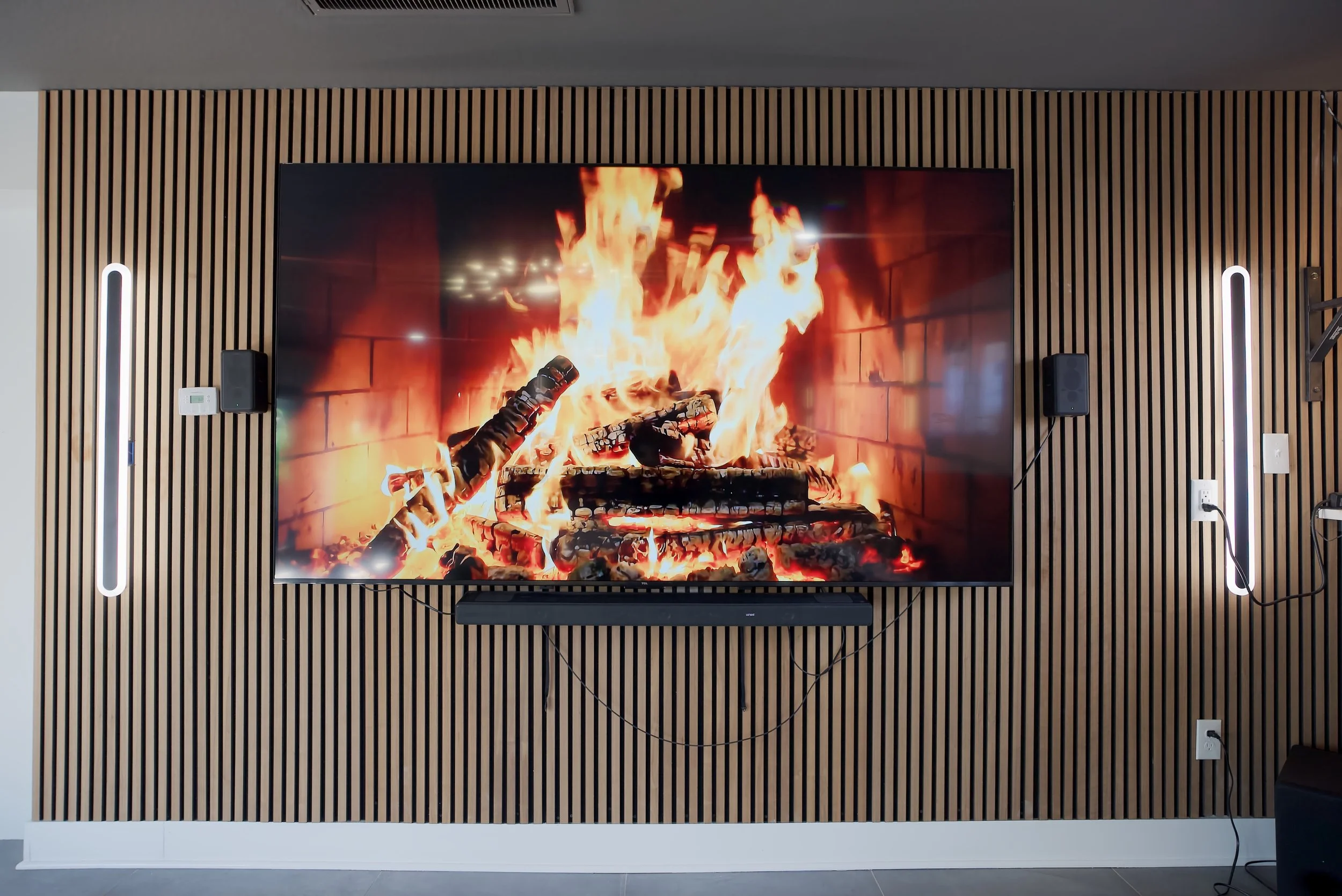 TV screen displaying a digital fire with logs and flames on a wood-paneled wall with speakers and lighting.