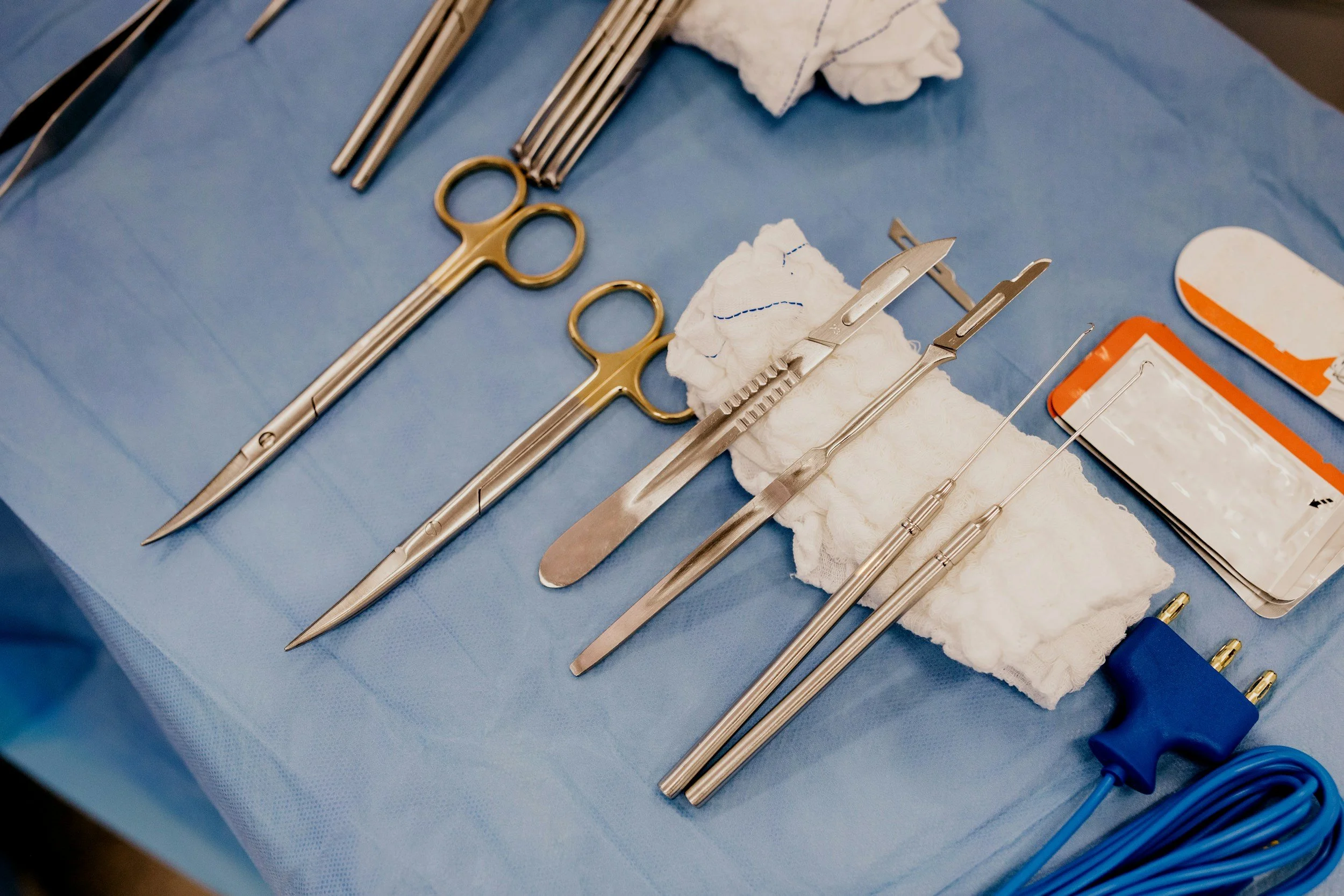 Medical surgical tools arranged on a blue sterile drape, including scissors, forceps, scalpels, and an electrocautery device.