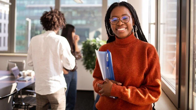 Jovem sorridente segurando pastas, em ambiente de escritório, com janelas e colegas ao fundo.