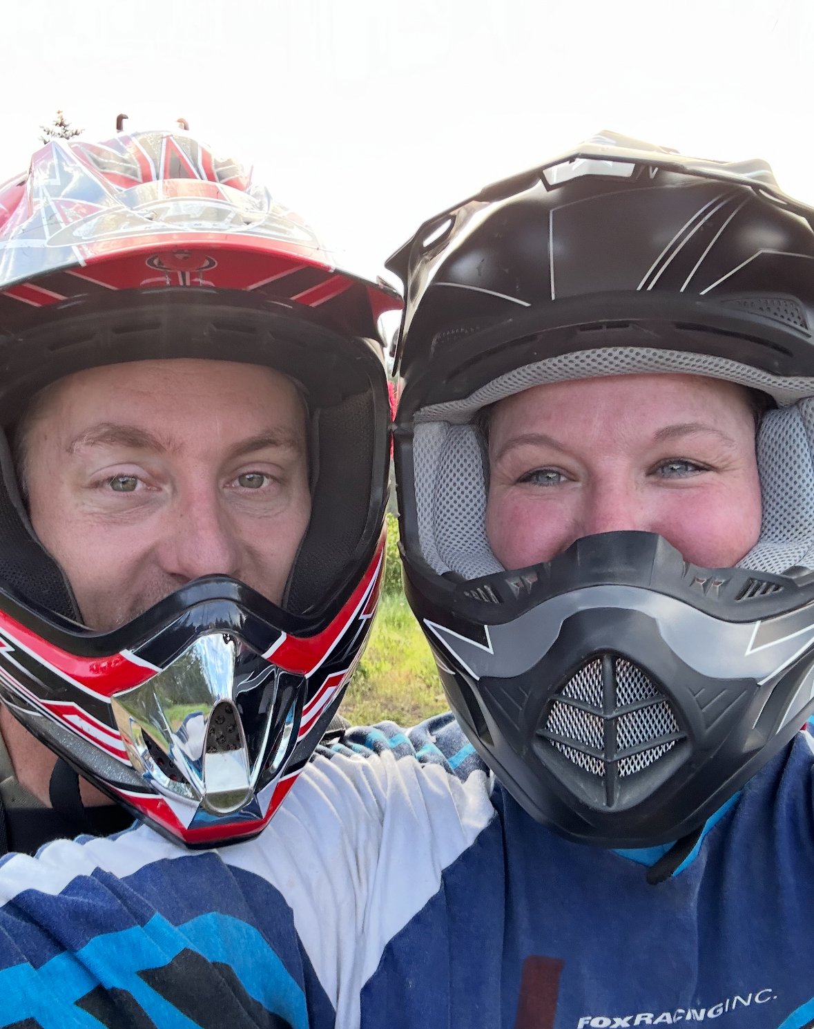 Two people wearing motocross helmets taking a close-up selfie outdoors.