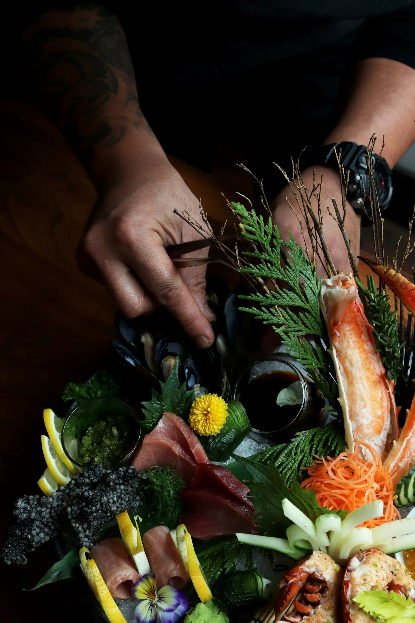 A person with a black watch arranging a seafood platter that includes lobster, sashimi, vegetables, lemon and lime slices, herbs, and condiments on a wooden table.