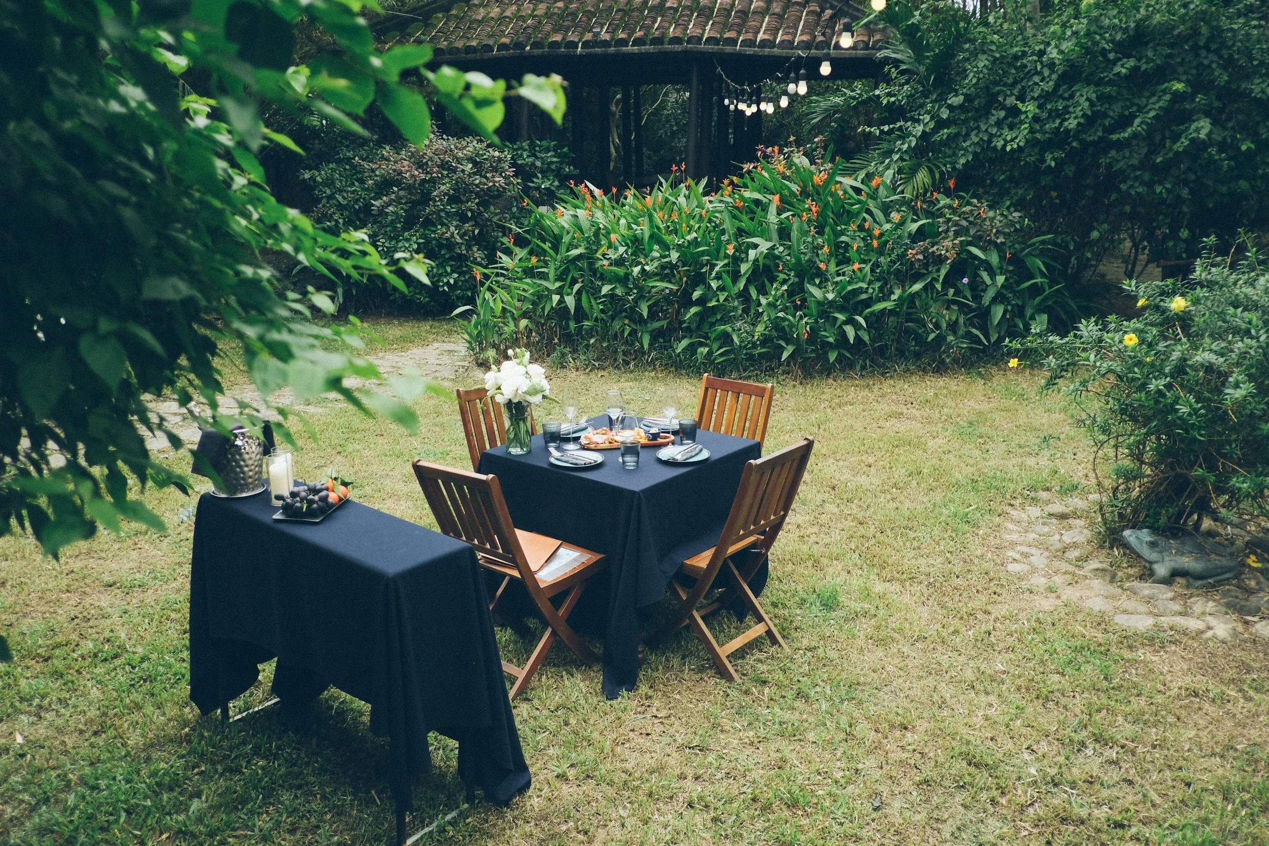 Outdoor garden setting with a table and four wooden chairs, set for a meal with plates, glasses, and a flower arrangement, surrounded by lush green bushes and plants.