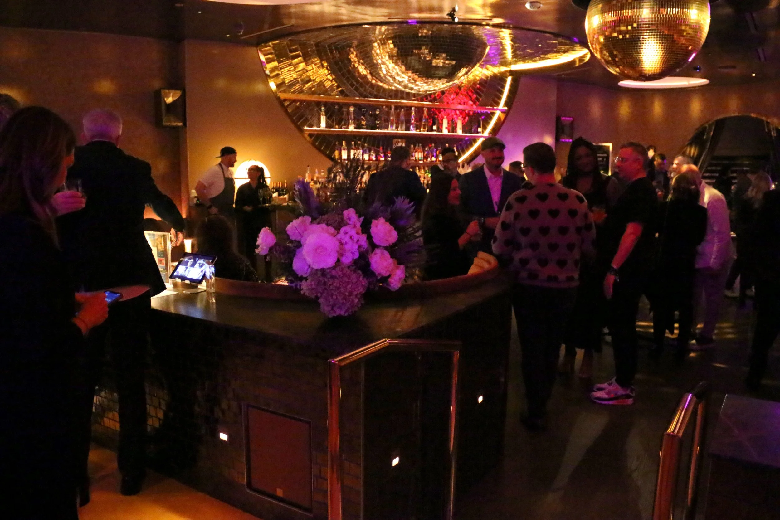 A lively bar with people socializing, a bartender behind the bar, and large decorative mirror and disco balls on the ceiling, illuminated by pink and purple lighting.