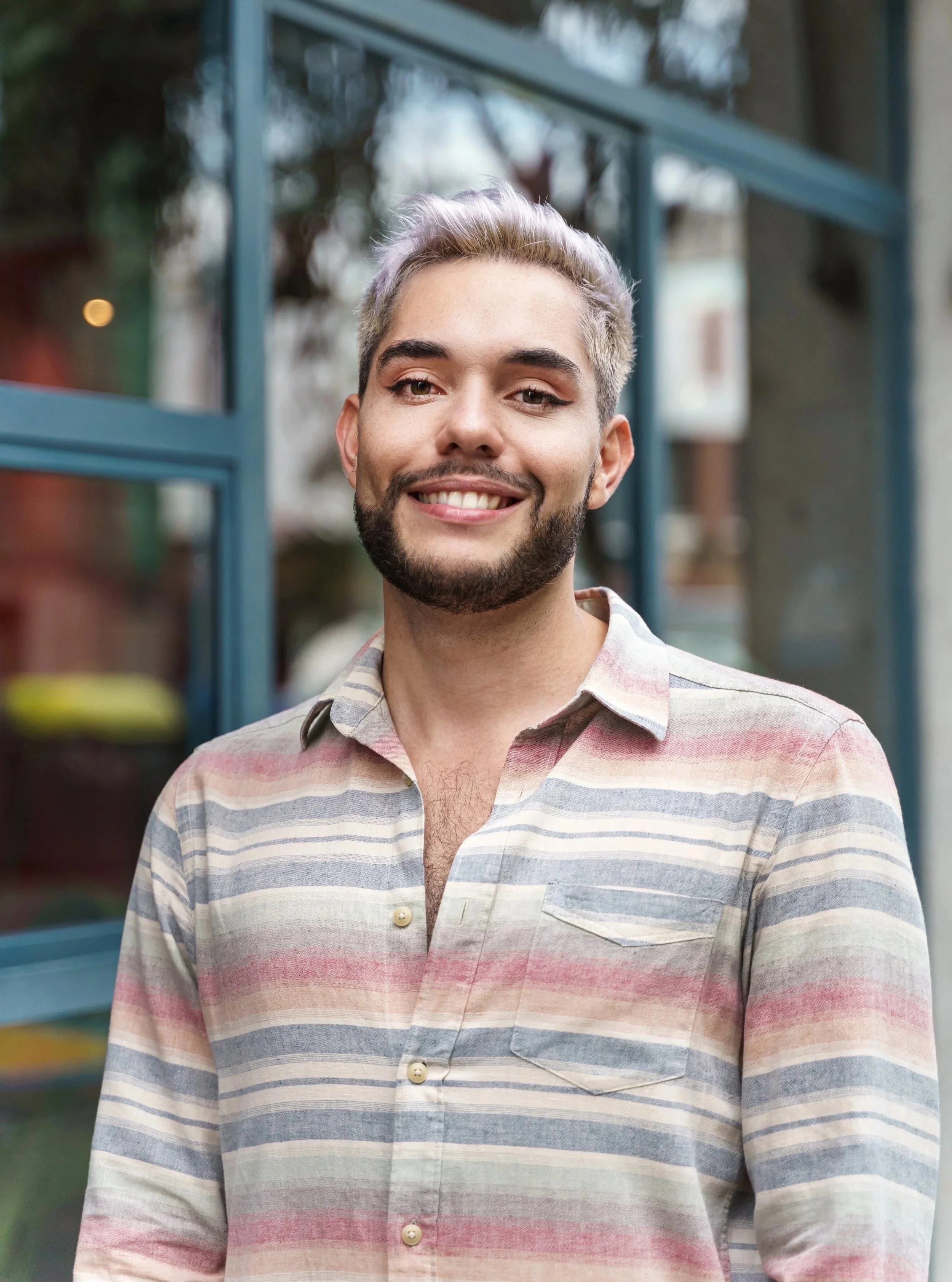 A young man with light hair and a beard, wearing a striped button-up shirt, is smiling outdoors in front of a building with glass windows.