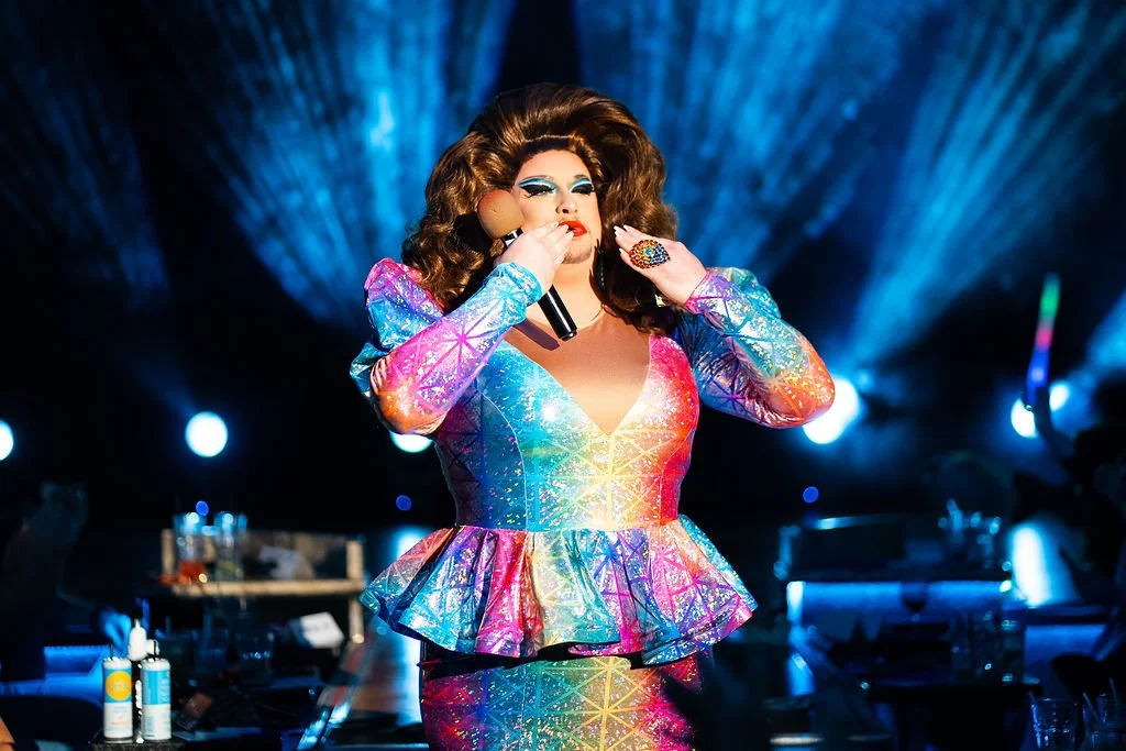 Miss LifeWalk 2025 Ginnifer with a G with voluminous hair in a colorful, shimmering outfit on stage with blue lighting and tables in the background.