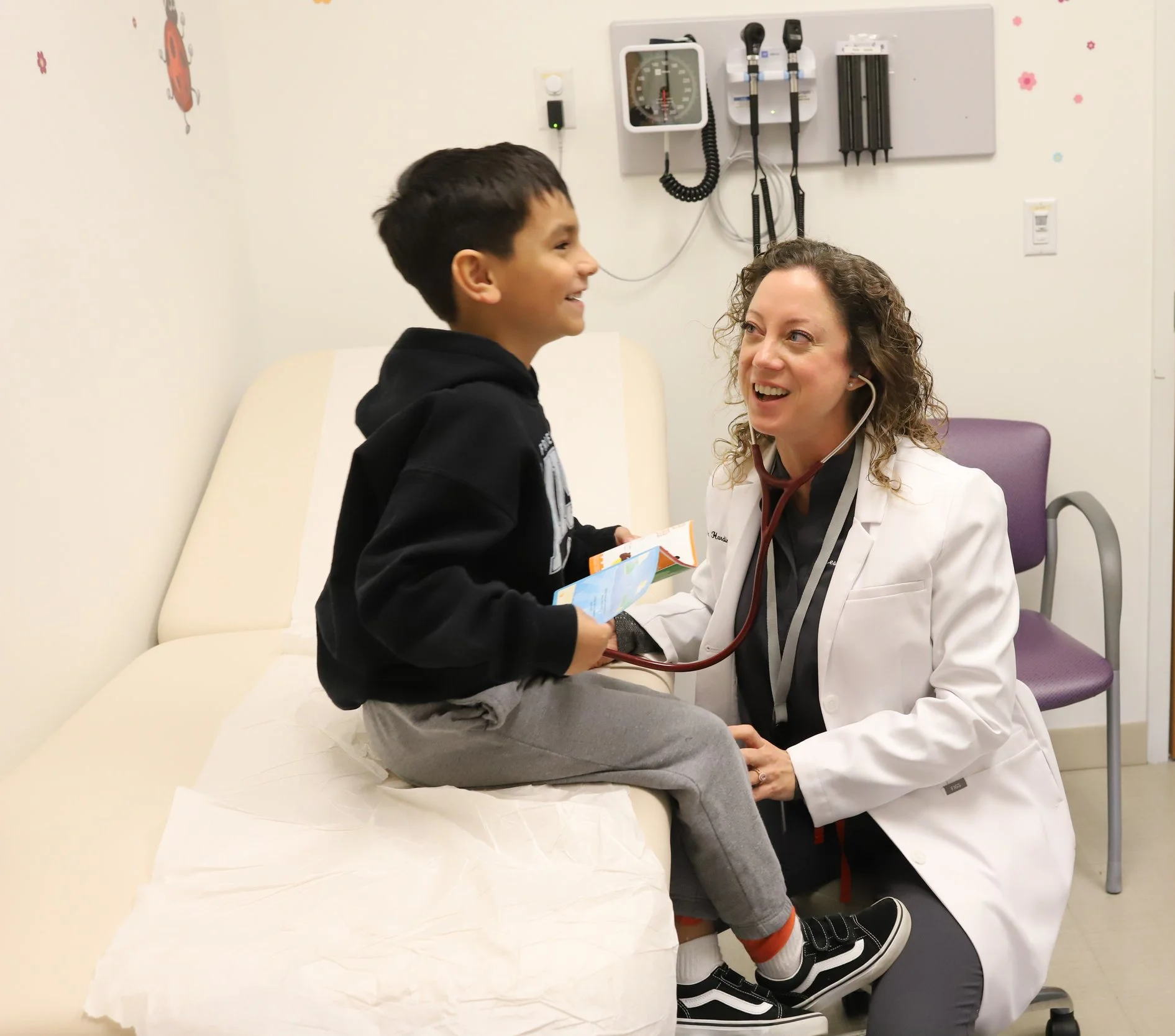 PHNTX Pediatric Provider Leslie Redden with young boy sitting on an examination table in a hospital room. The boy is smiling and holding a book, with colorful wall decorations present.