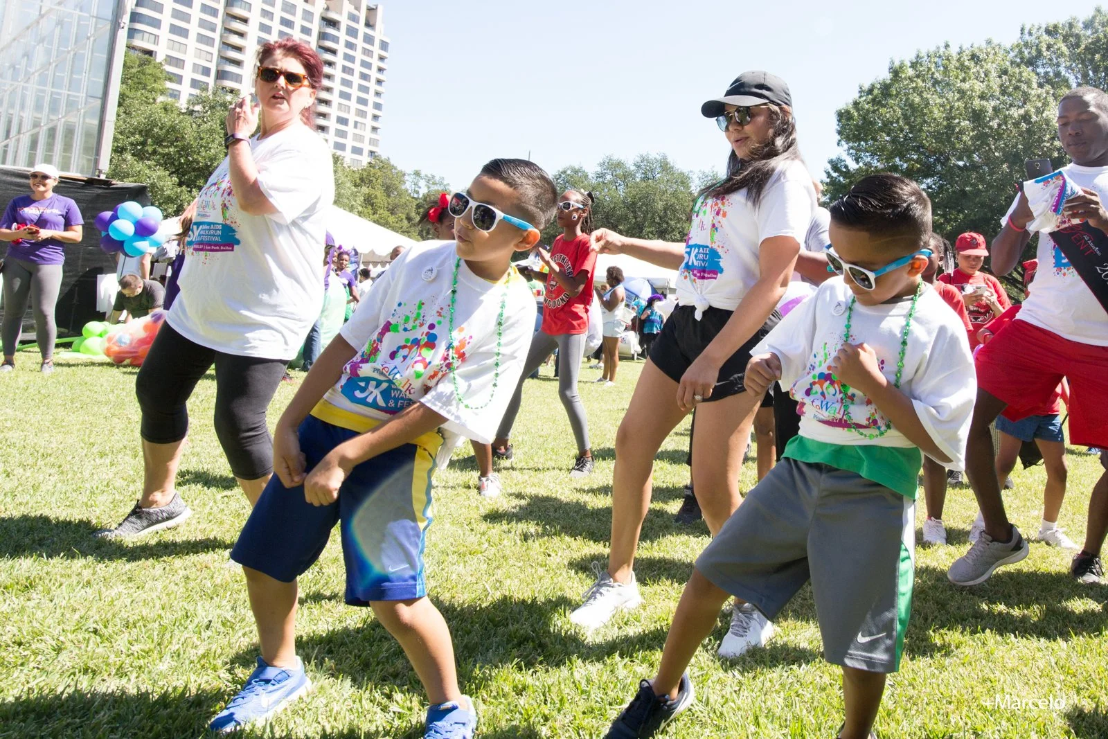 Group of children and adults dancing on grass at an outdoor event, with tents, balloons, and tall buildings in the background.
