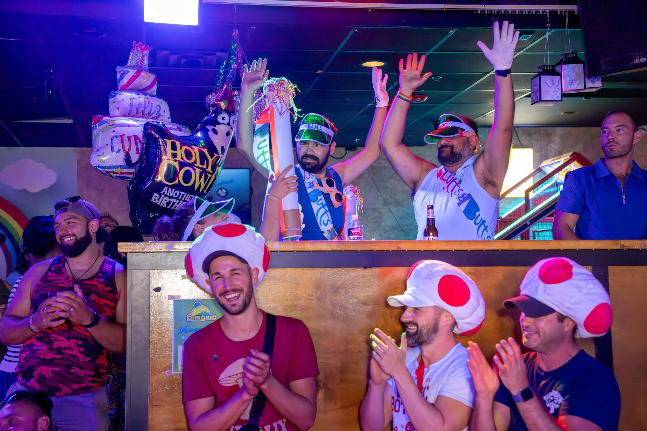 People celebrating with balloons, hats, and costumes themed around Mario. Some are wearing mushroom hats, others are in T-shirts and sunglasses, with colorful balloons and decorations in the background.