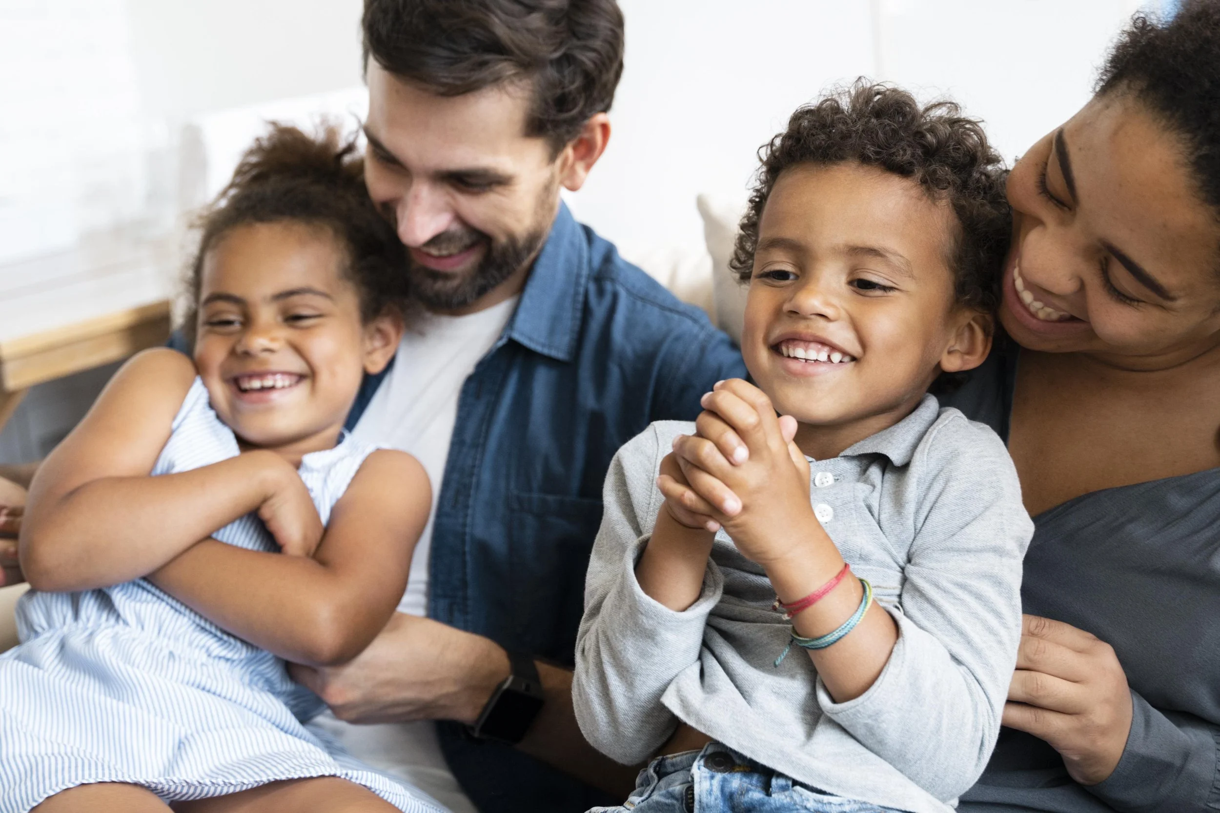 A happy family of four, including two children and two adults, sitting together and smiling in a cozy home environment.