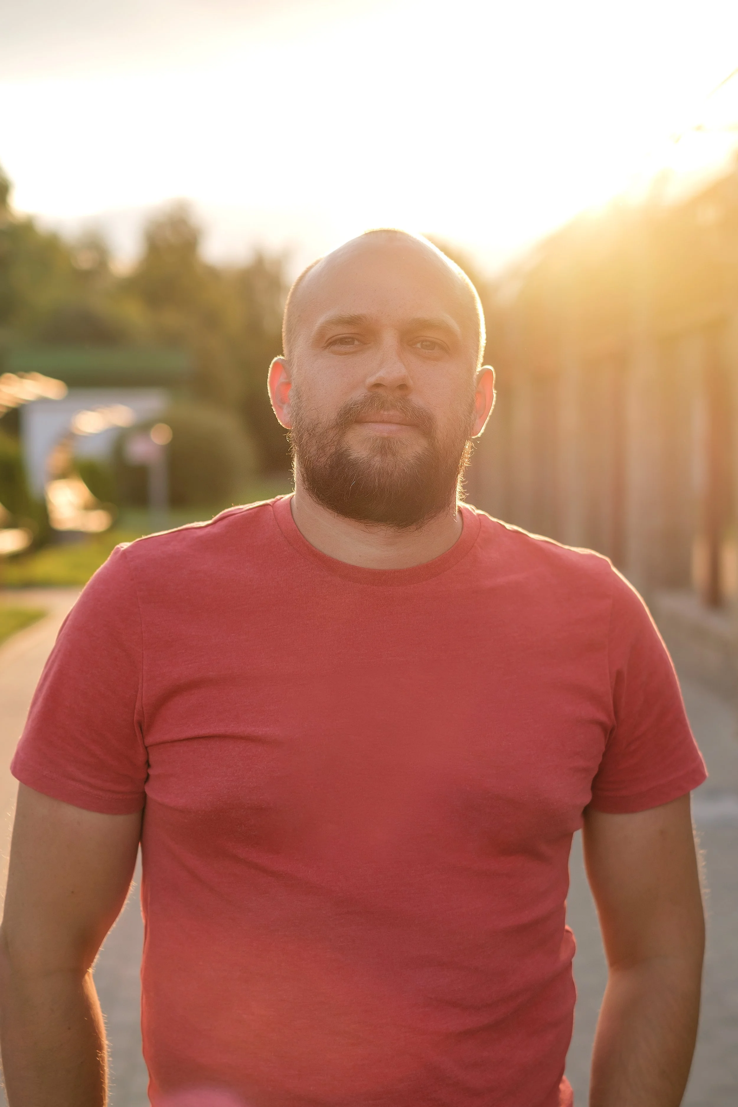 A man with a beard and a red t-shirt standing outdoors during sunset with sunlight backlighting him.