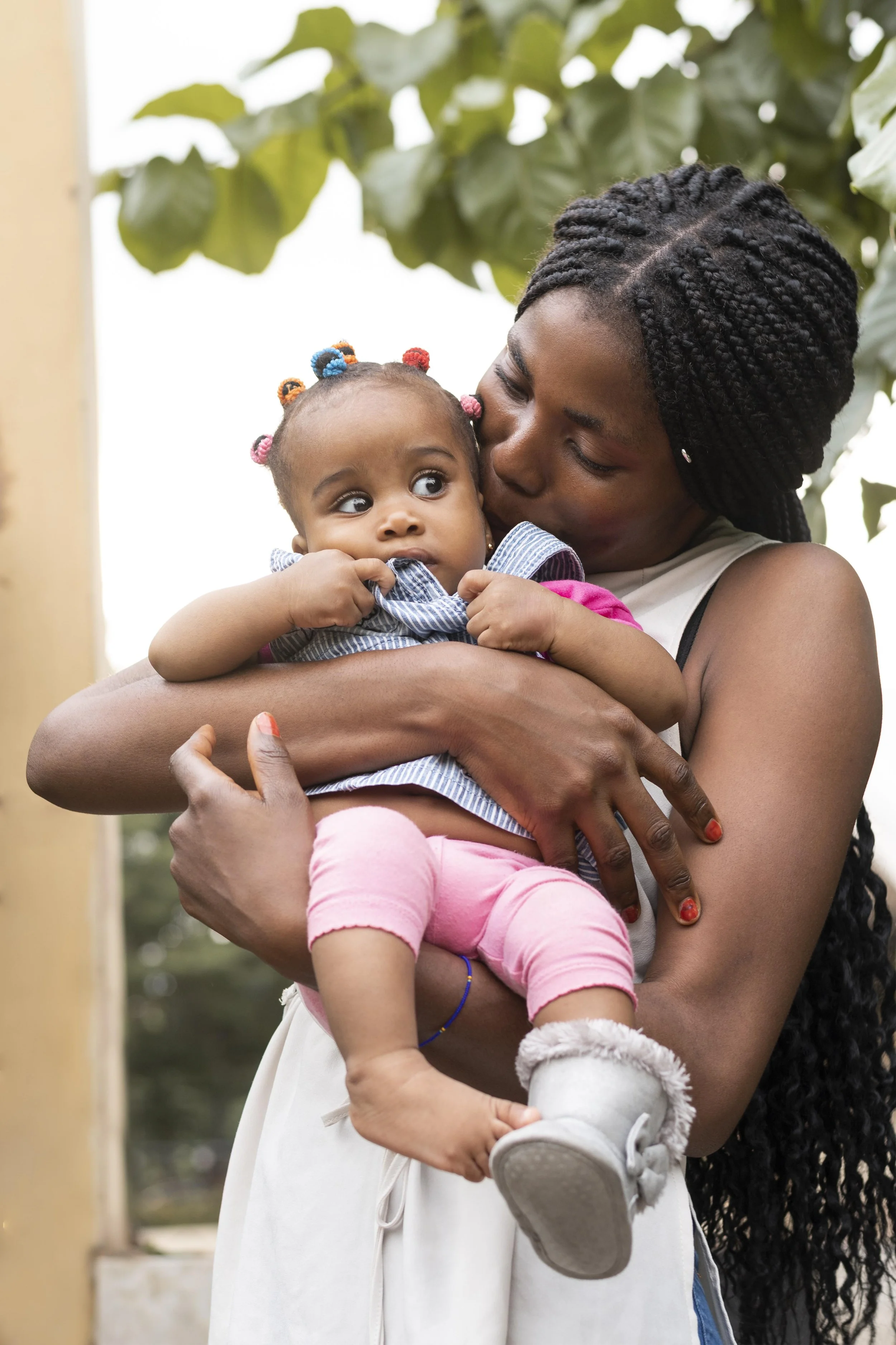 A woman holding a young girl. The woman is kissing the girl on the cheek, and the girl is looking to the side. They are outdoors with green leaves in the background.