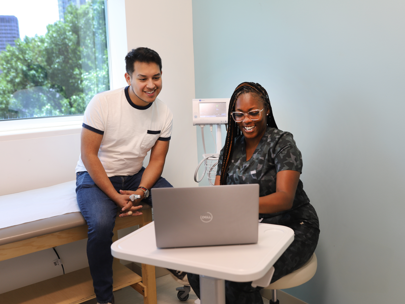 A PHNTX healthcare professional and a young man looking at a laptop together in a medical office, with a window showing trees outside and medical equipment on the wall.