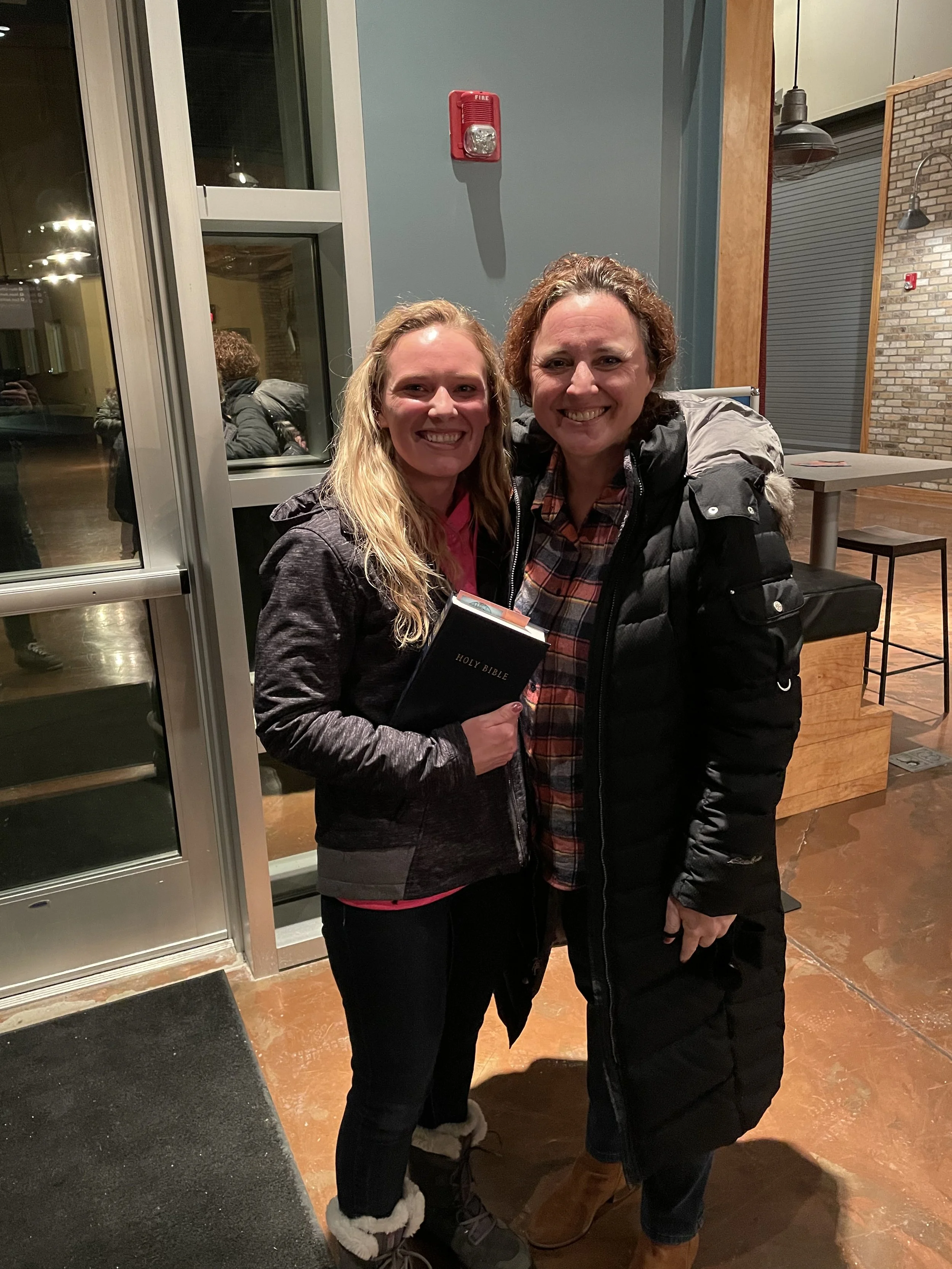 Two women smiling and posing inside a building near a door, one holding a Holy Bible, wearing winter clothing.