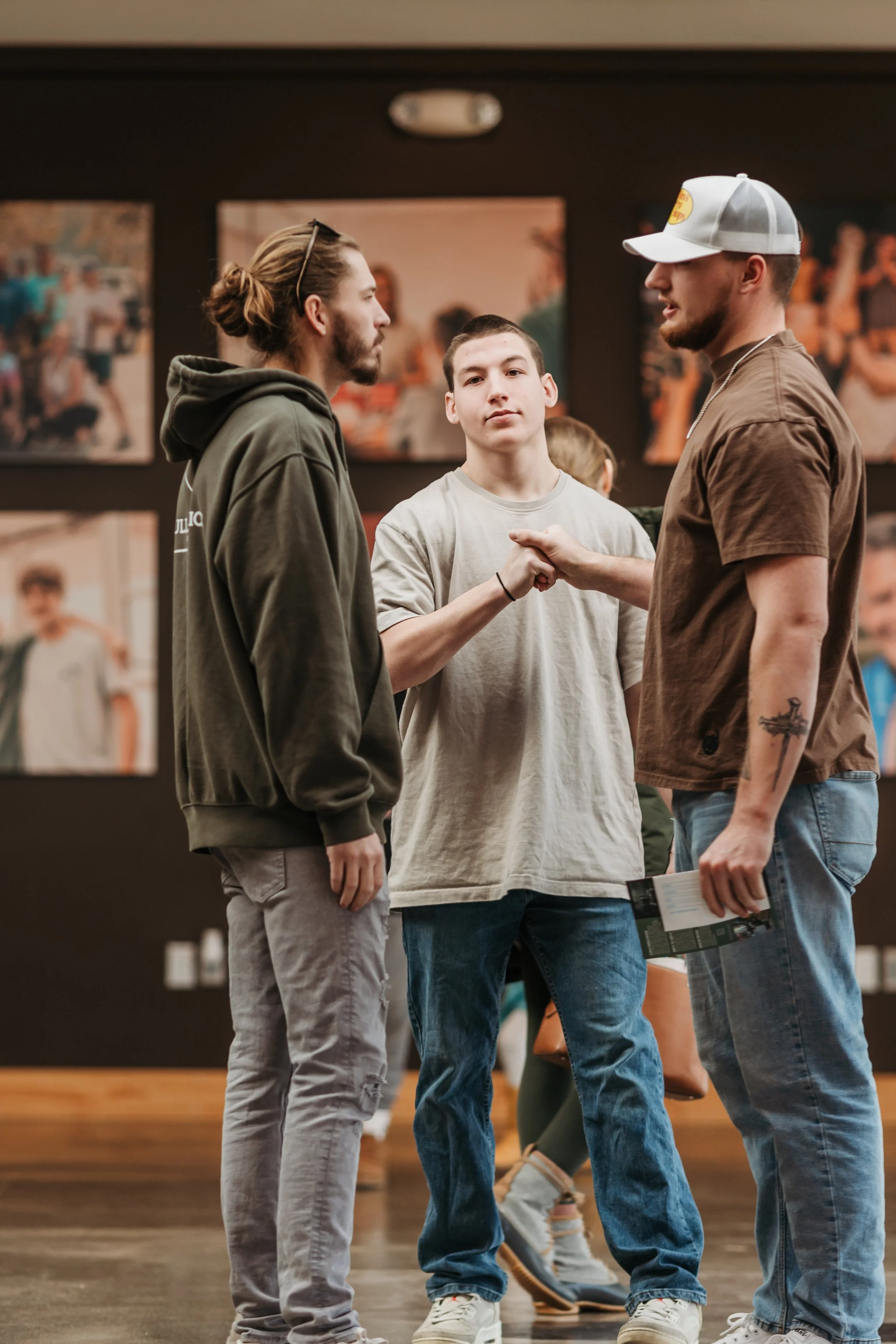 Three young men engaged in conversation in an indoor setting with framed pictures on the wall in the background.