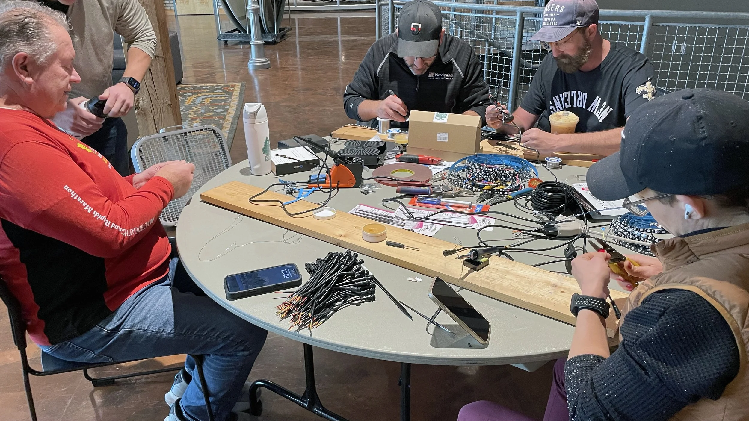 Group of people working on electronics projects at a round table, with wires, tools, and electronic components scattered across the surface.