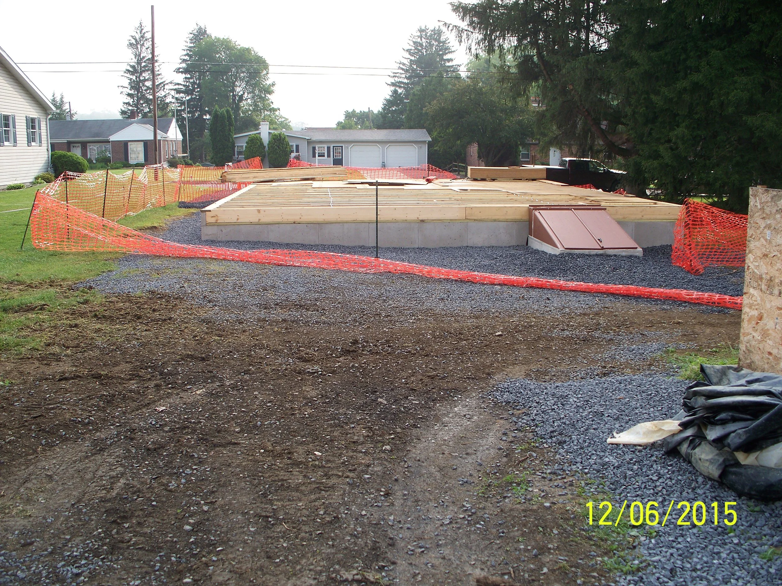 Construction site with a foundation being laid, surrounded by orange safety fence and gravel, in a residential neighborhood with trees and houses in the background.
