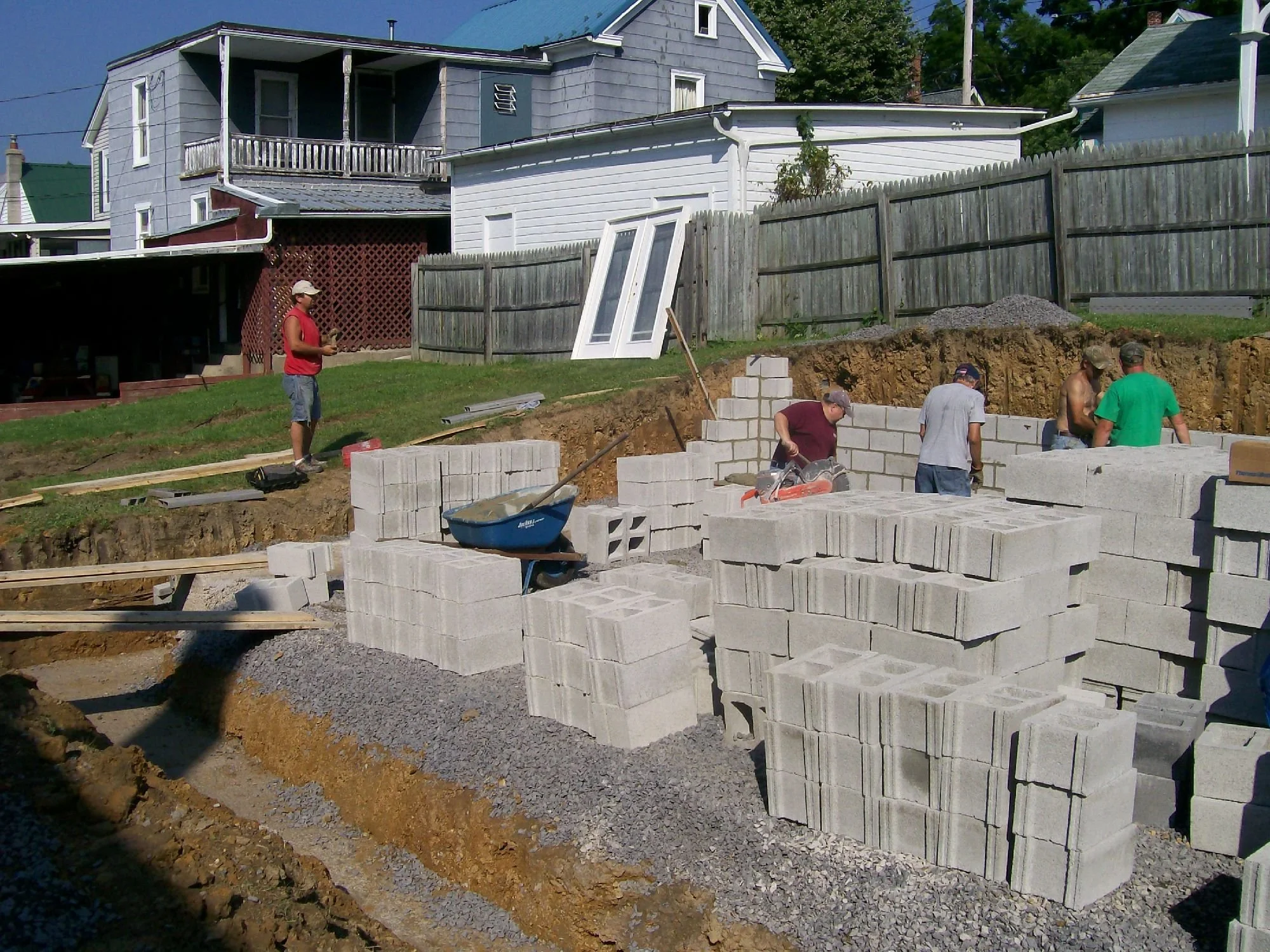 Construction workers building a brick wall on a residential site, with stacks of concrete blocks, a wheelbarrow, and construction tools visible in the foreground. A person in a red shirt and cap is standing on a grassy area nearby.