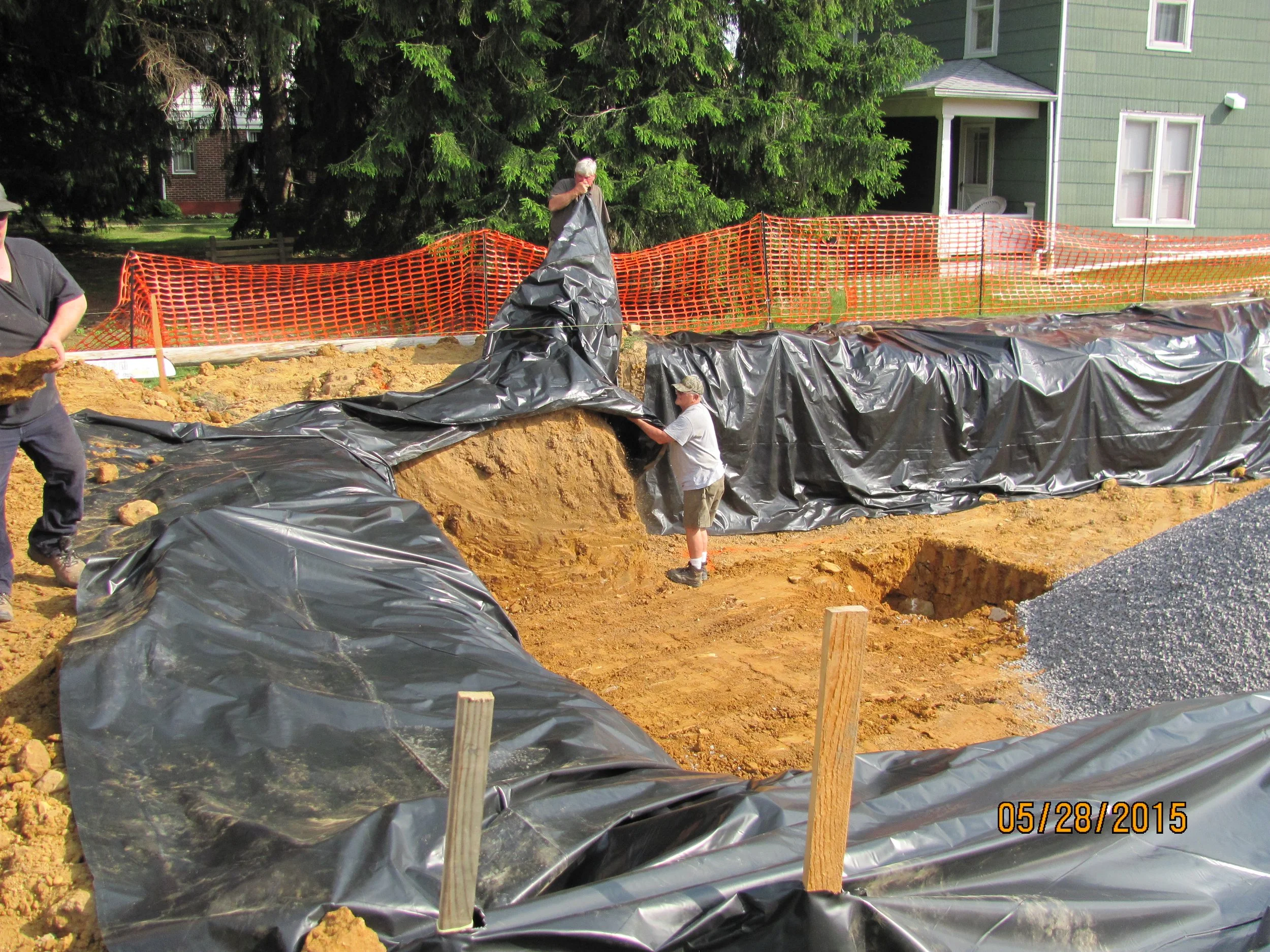 People working on a construction site with black plastic sheeting and orange safety fencing, preparing the ground for a foundation or pool.