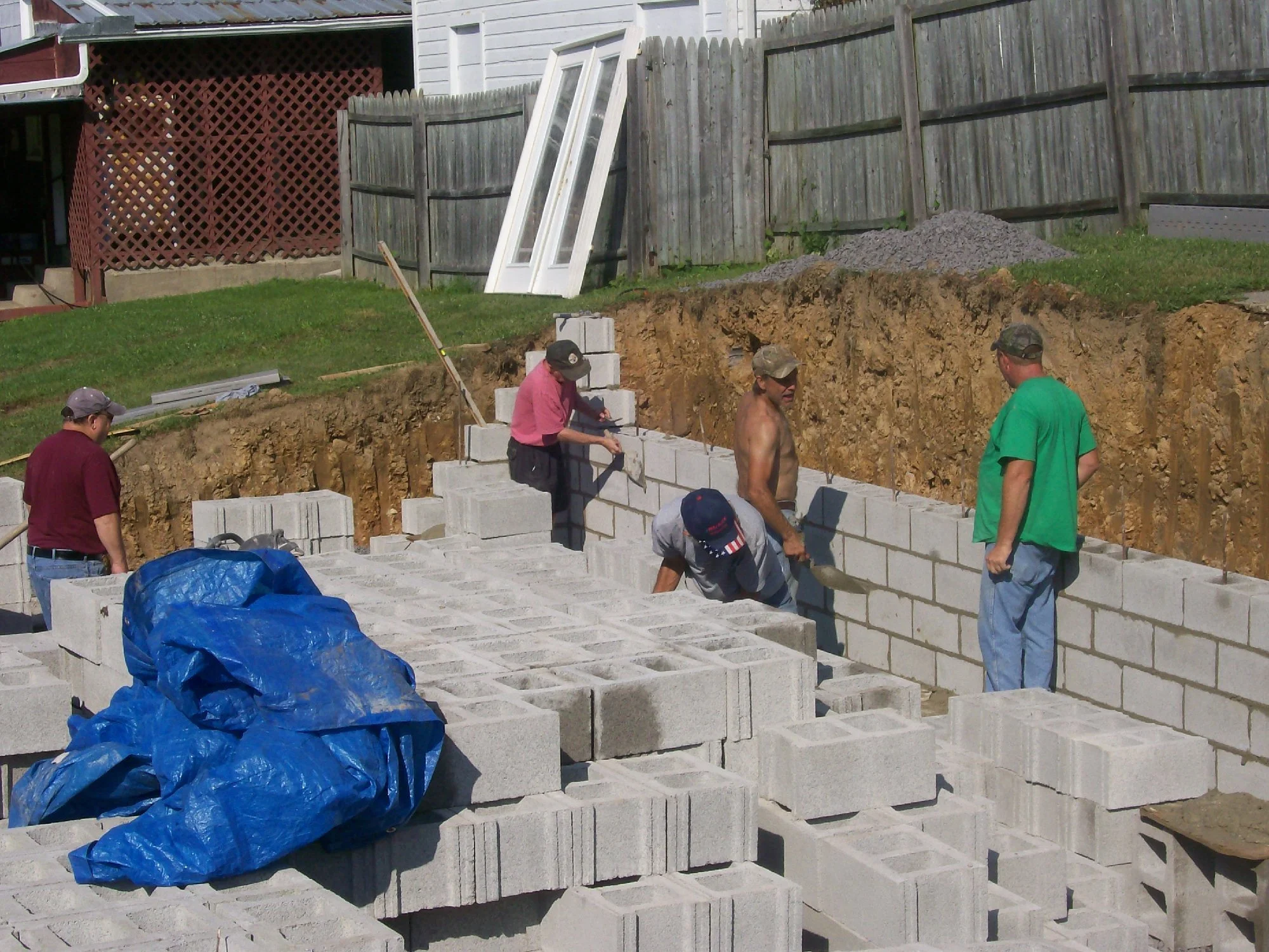 Workers laying concrete blocks to build a wall on a construction site, with some workers wearing casual clothing and others shirtless.