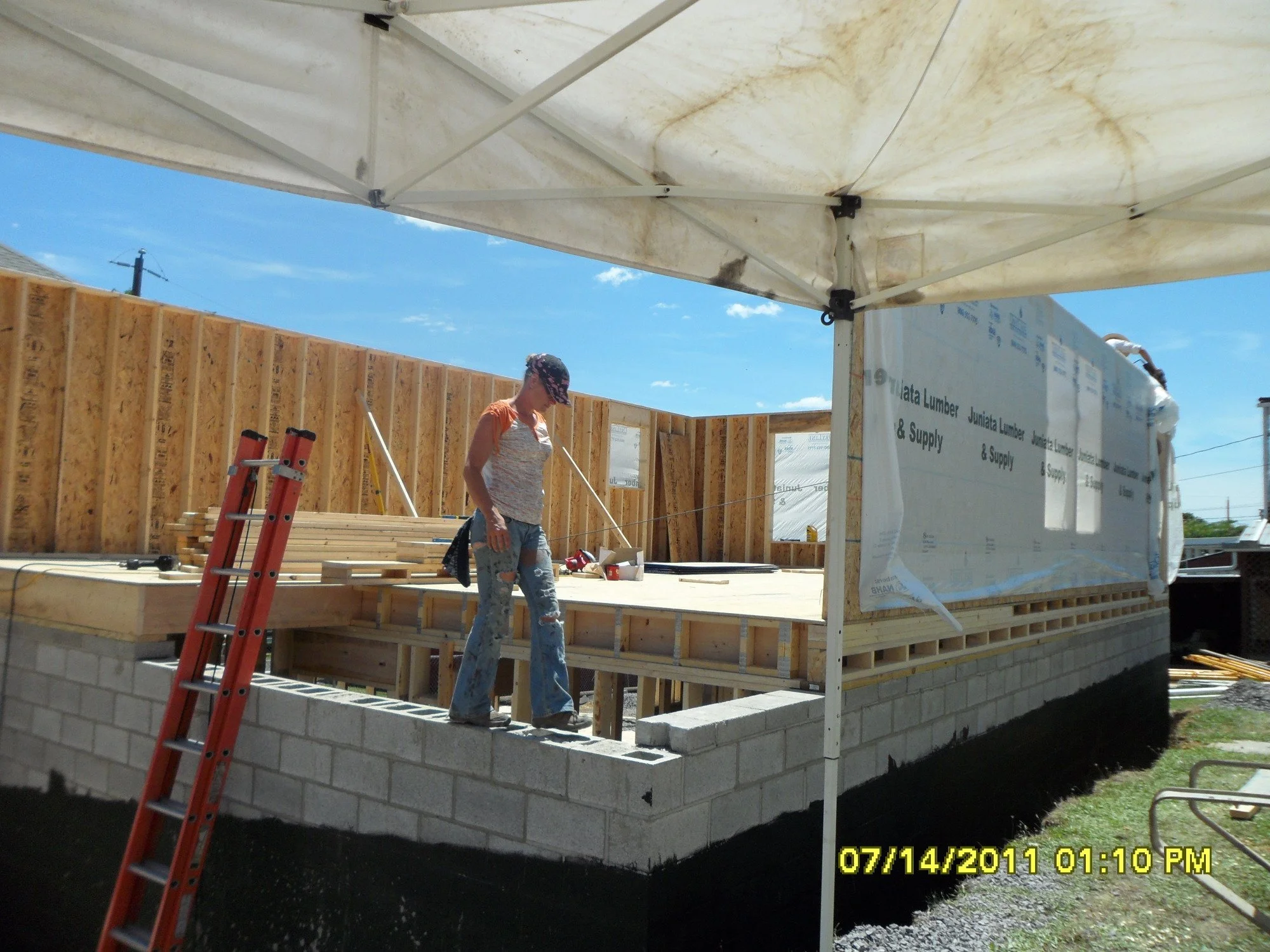 A woman working on a construction site under a canopy, with wooden framing and a concrete block foundation visible.
