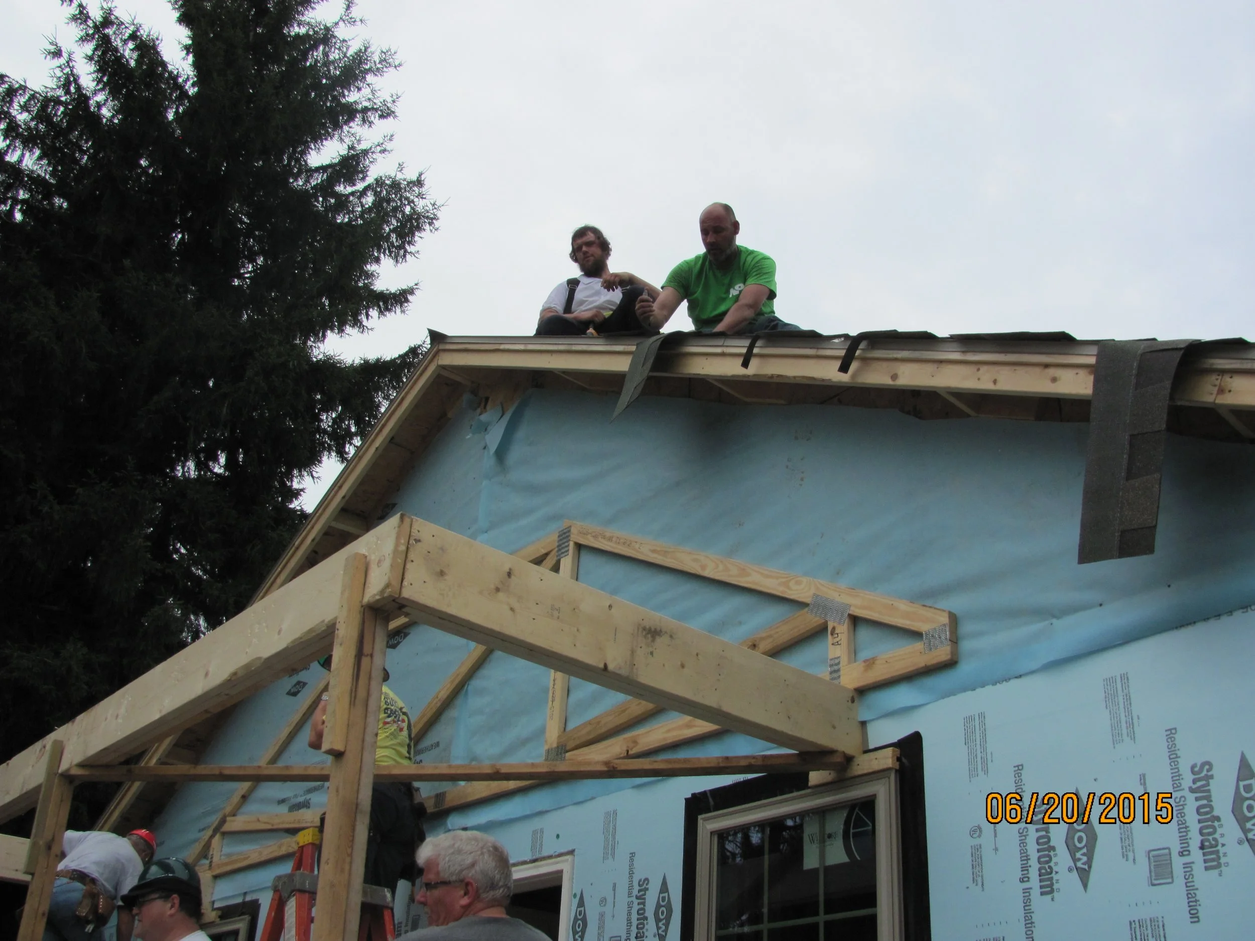 Three men working on the roof of a house under construction, with two men sitting on the roof and one man standing below. The house is partially covered with blue insulation material, and a wooden frame is being built in front of the house. The date 