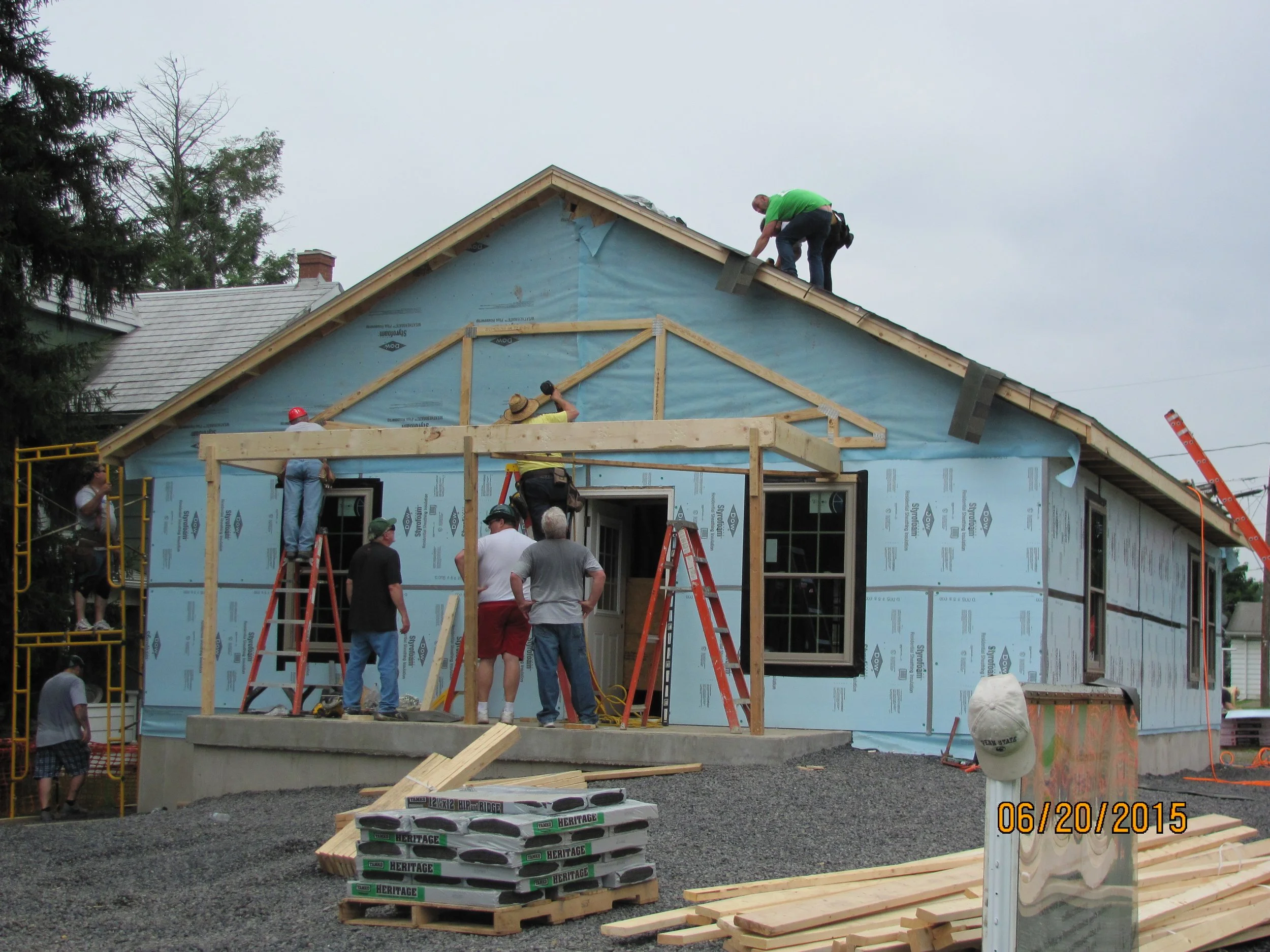 Workers building a house, some on the roof and some on the ground, with construction materials and scaffolding present.