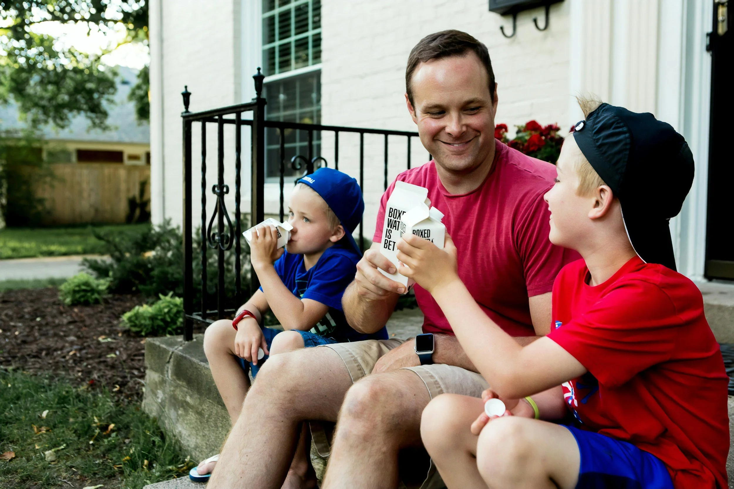 Father and two young boys sitting on a porch steps, drinking milk from cartons. The father is smiling and holding a carton while the boys, wearing hats, drink from their cartons.