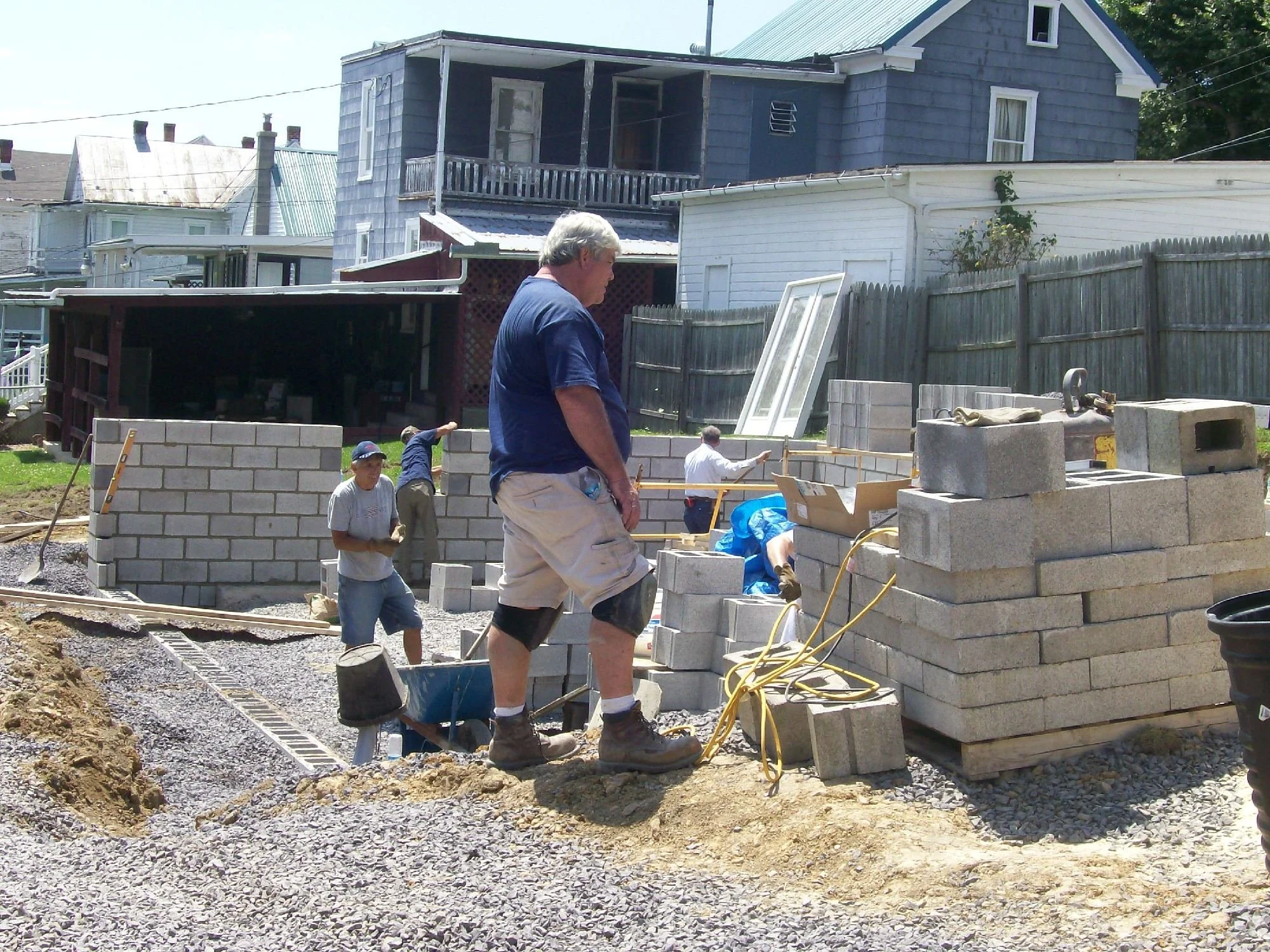 Construction workers building a brick wall outdoors on a sunny day, with a blue house and a white fence in the background.