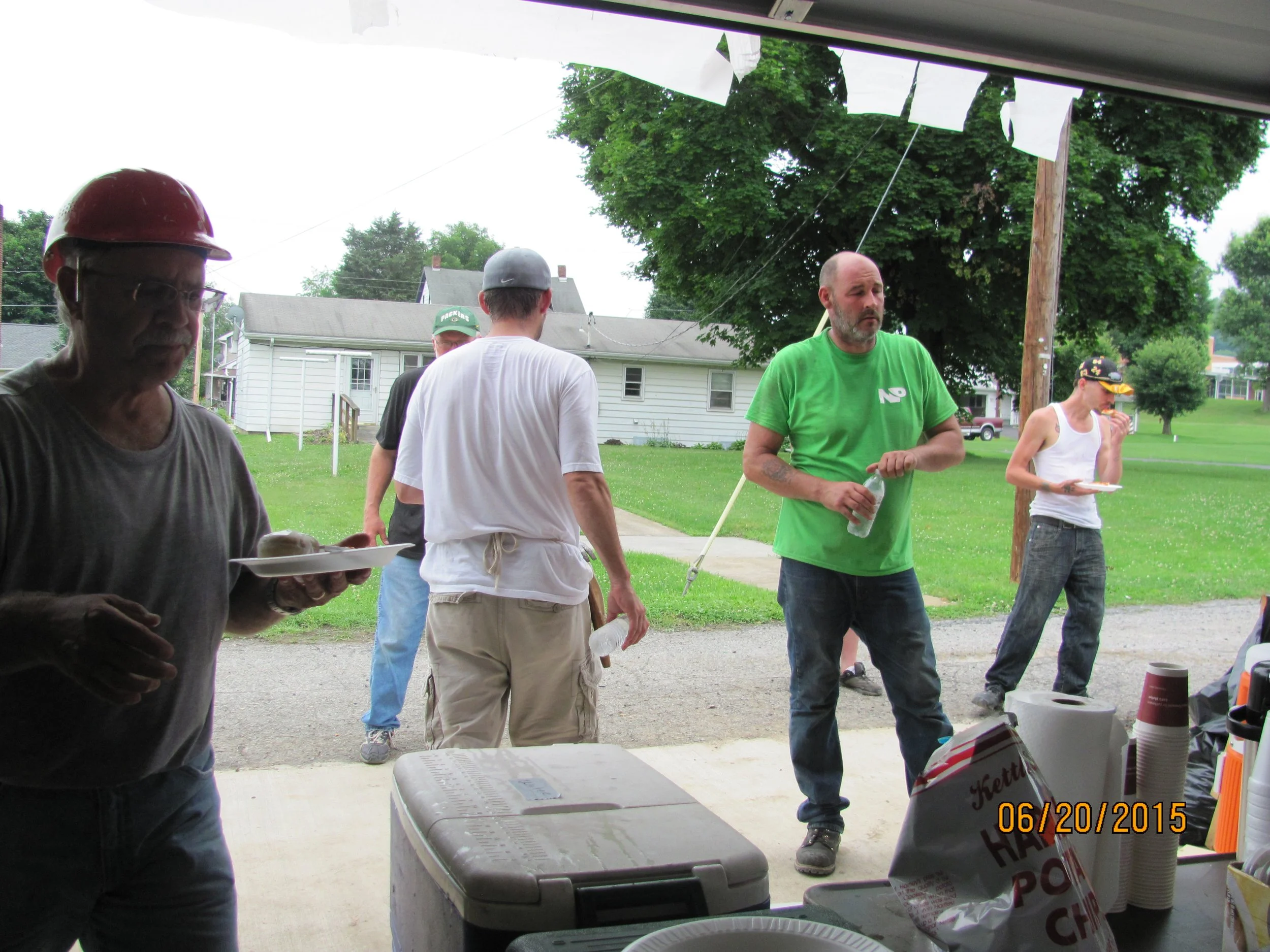 Group of people gathered outside under a porch, some eating, with a grassy yard and houses in the background, date stamped June 20, 2015.