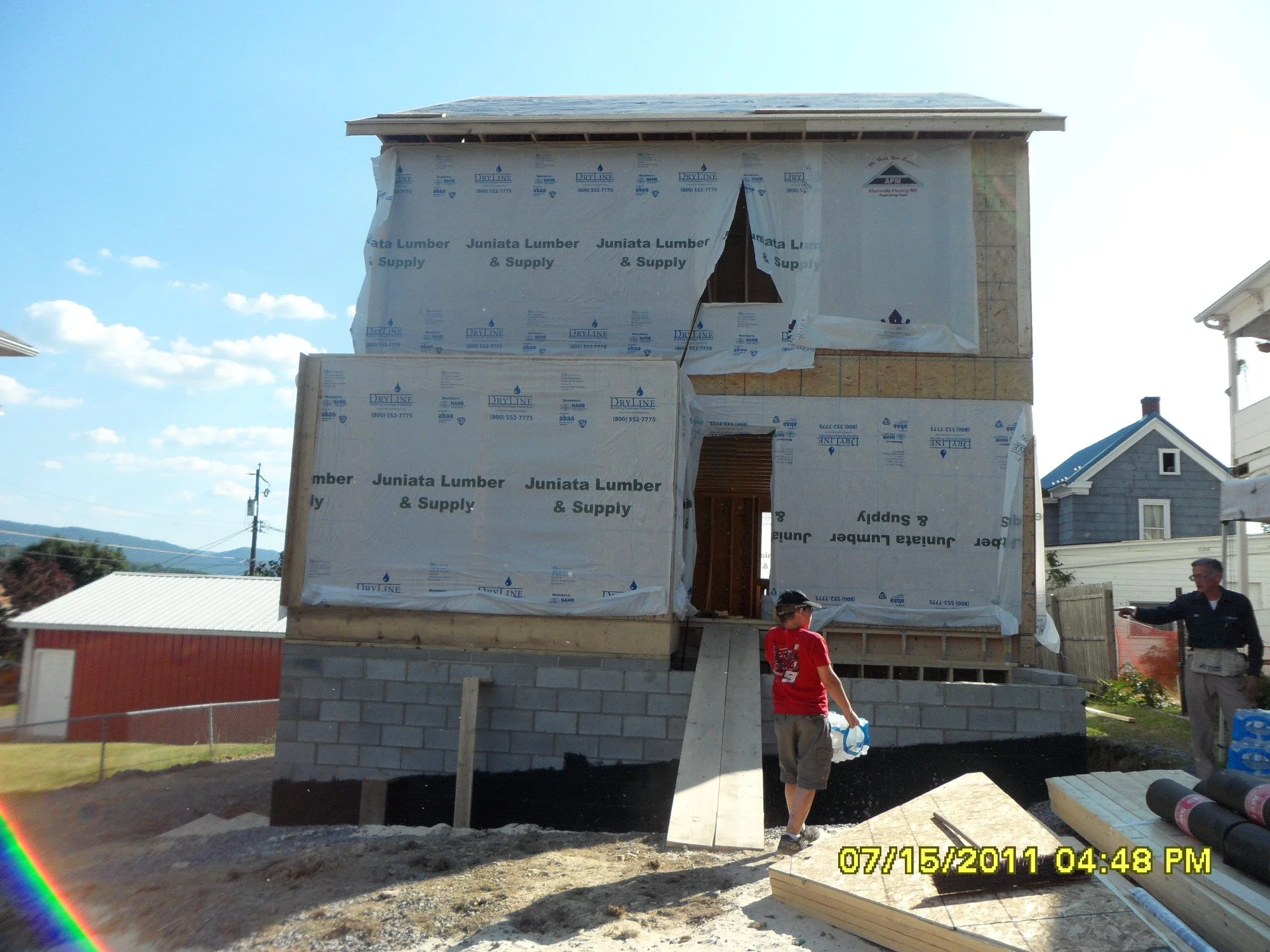 Two-story house under construction with wooden framing and insulation wrapping, construction workers present, and neighboring houses visible in the background.
