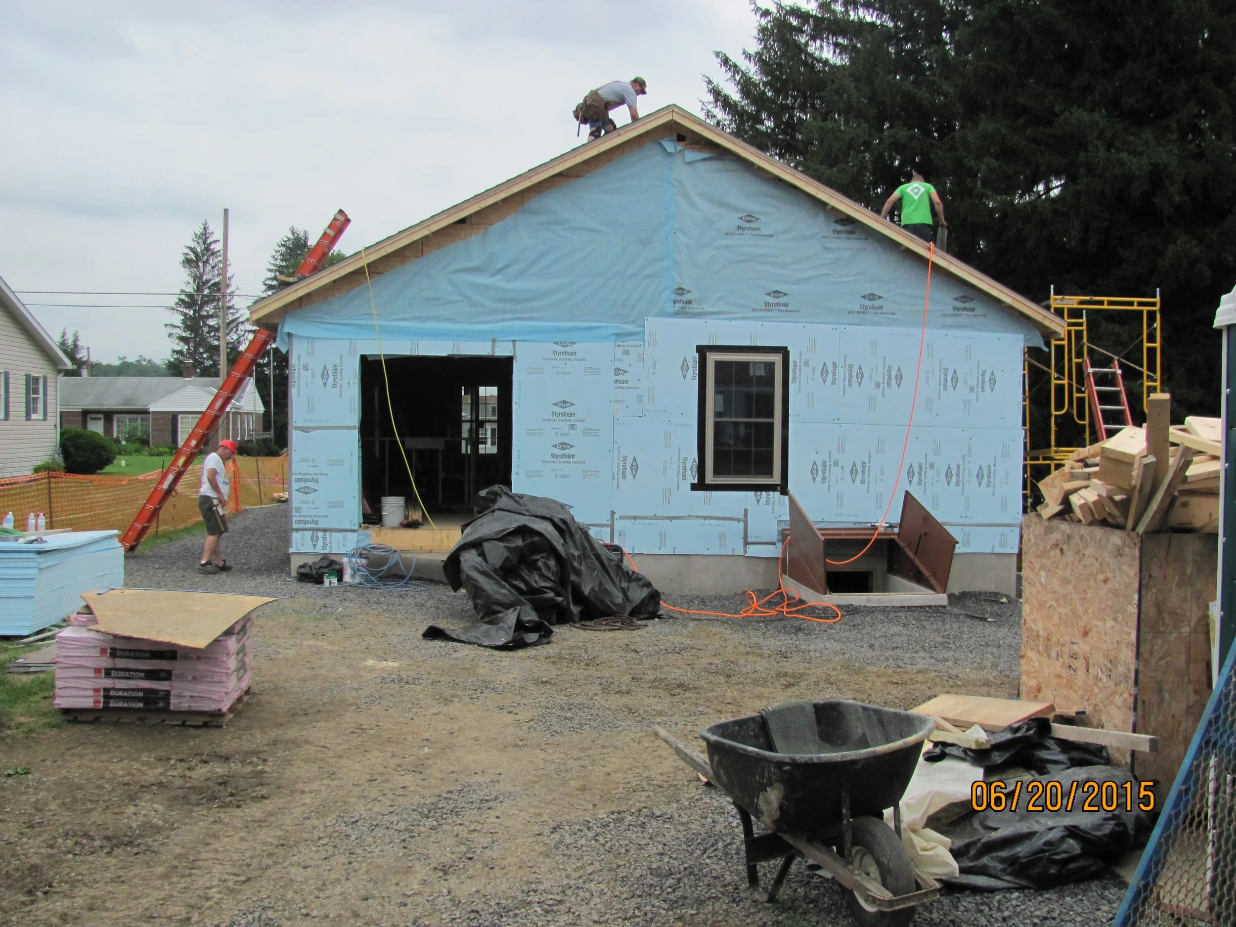 Construction workers are building a house, with one on the roof and another on a scaffold, while a third worker walks nearby. Construction materials and tools are scattered around the site, including a wheelbarrow, lumber, insulation, and a black tar