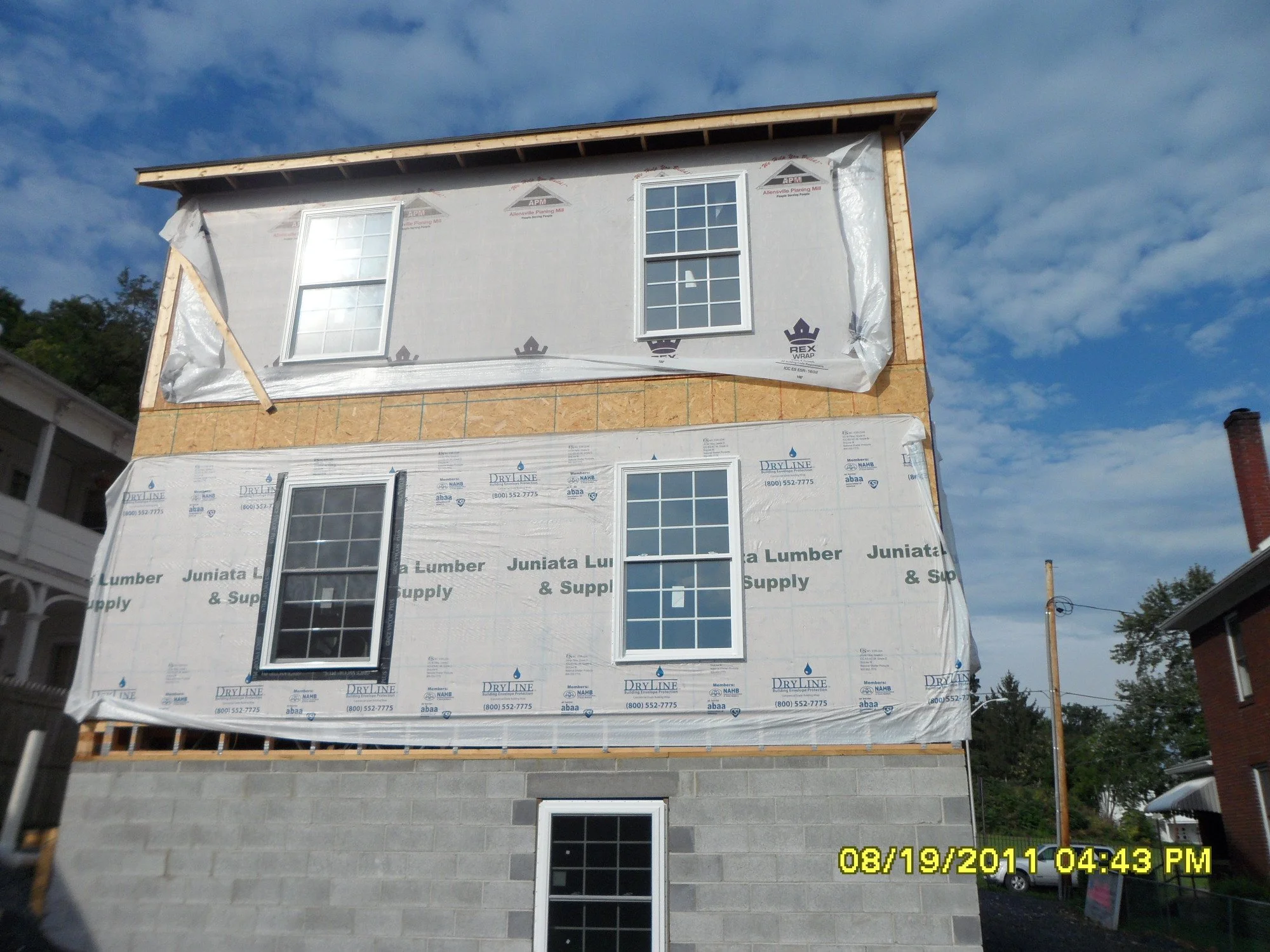 A three-story house under construction with windows installed on the upper two floors. The building has a concrete block foundation, and the middle and top sections are wrapped in construction paper, while the upper section is covered with a weather-