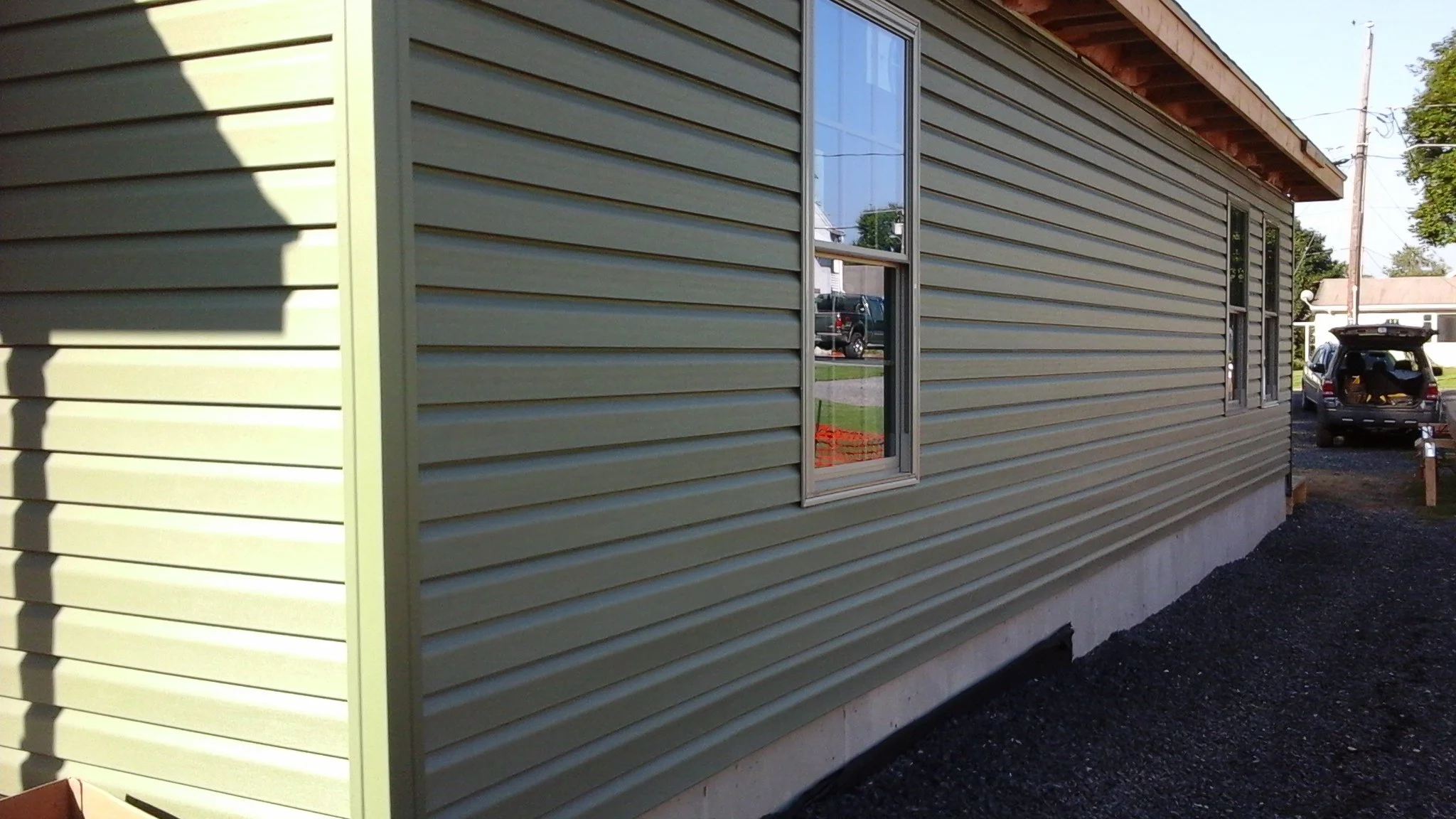 Side view of a house with beige horizontal vinyl siding, two windows, and a concrete foundation. There is a gravel path along the house, and a black SUV with open trunk parked nearby.