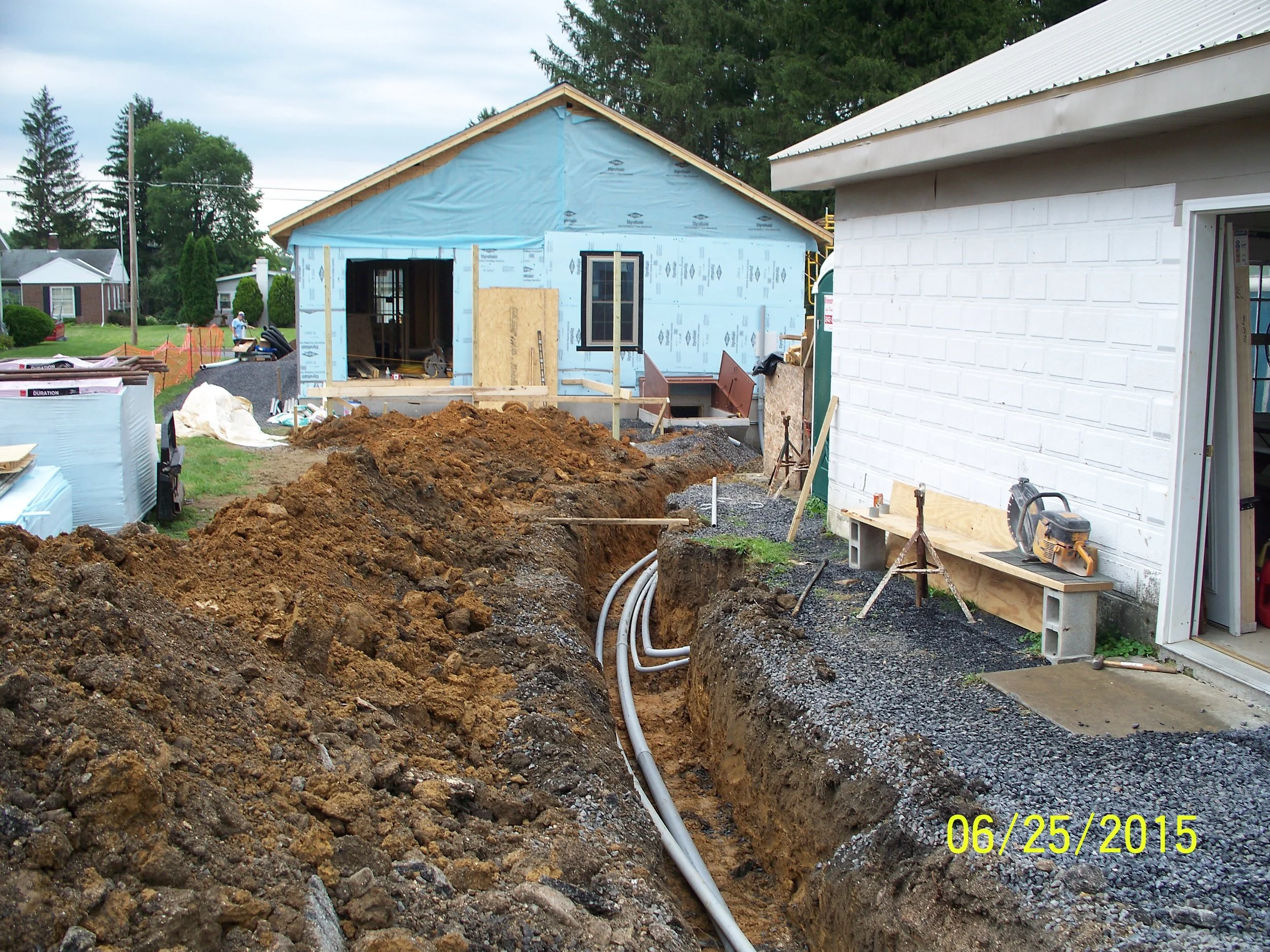 Construction site featuring a trench with electrical conduit pipes, a partially built house in the background, and a white brick building on the right. The date on the photo is June 25, 2015.