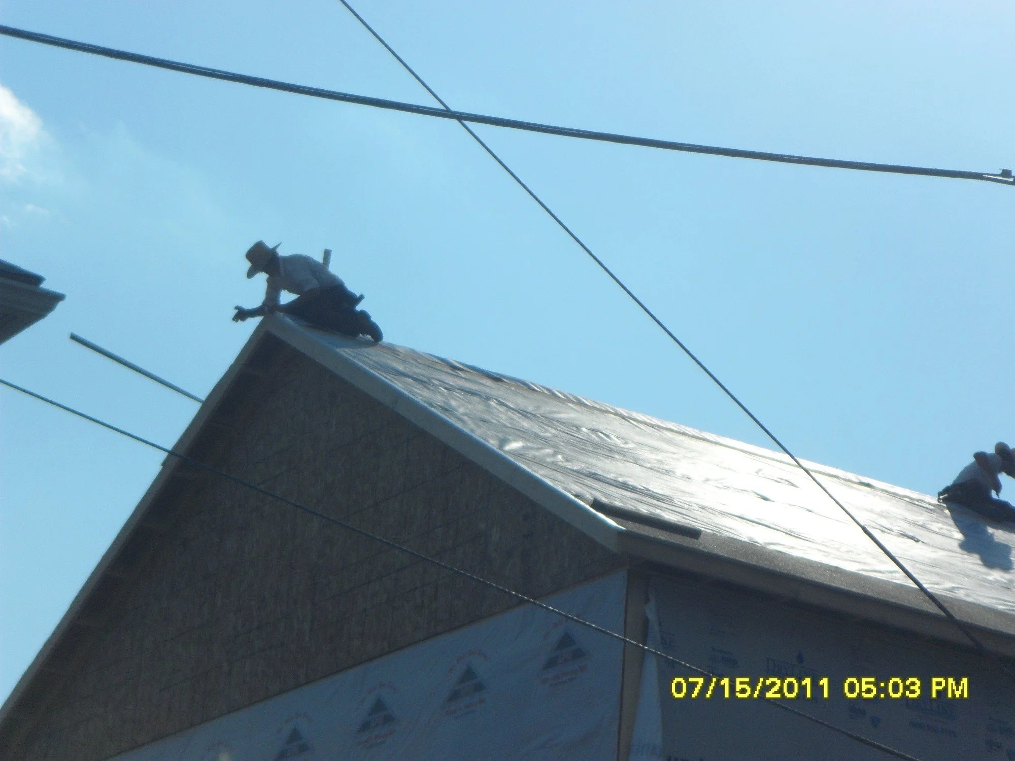 Two workers on the roof of a building installing or repairing the shingles under a clear blue sky with power lines overhead.