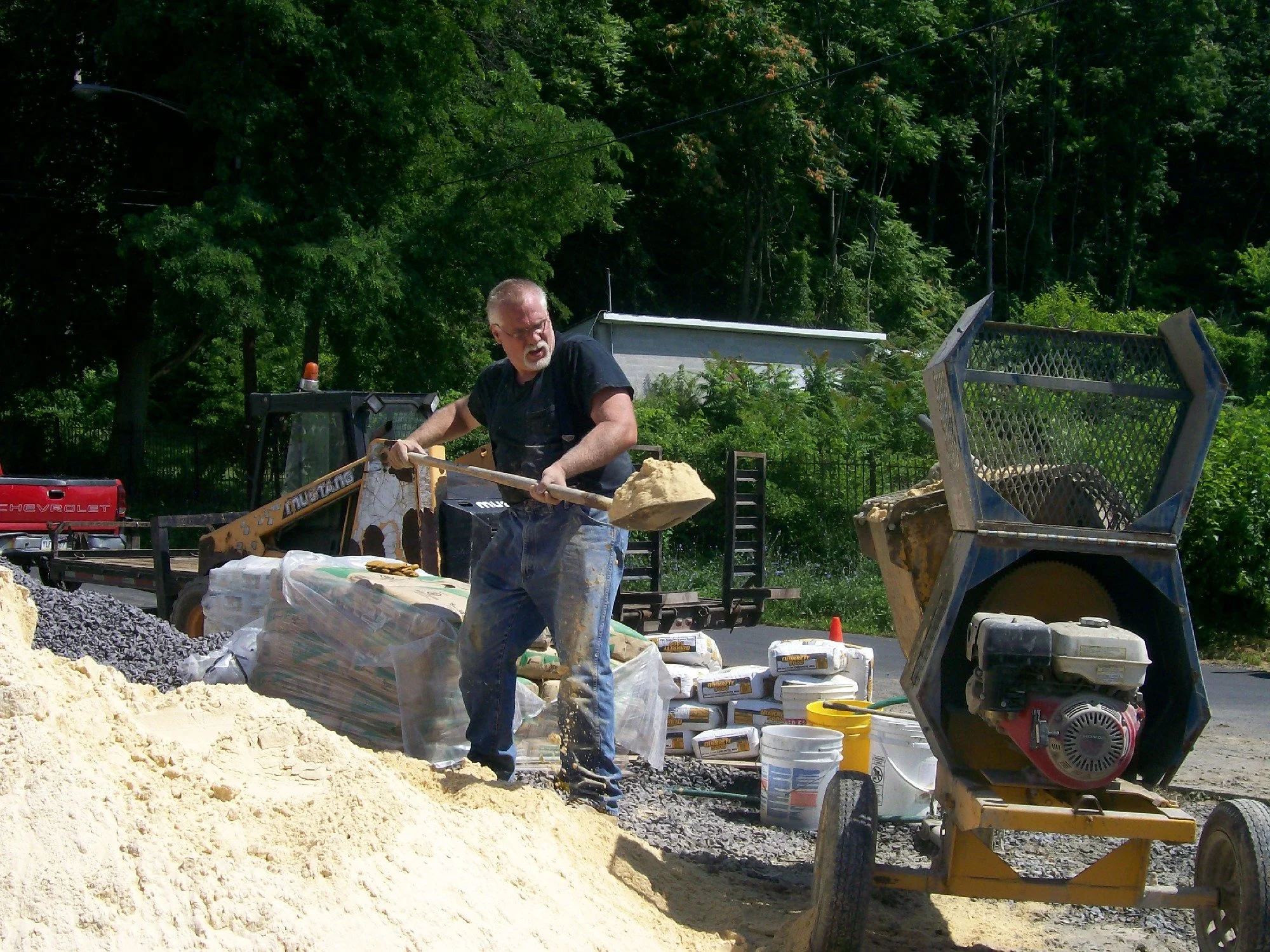A man working outdoors with construction equipment, shoveling sand on a construction site with trees in the background.
