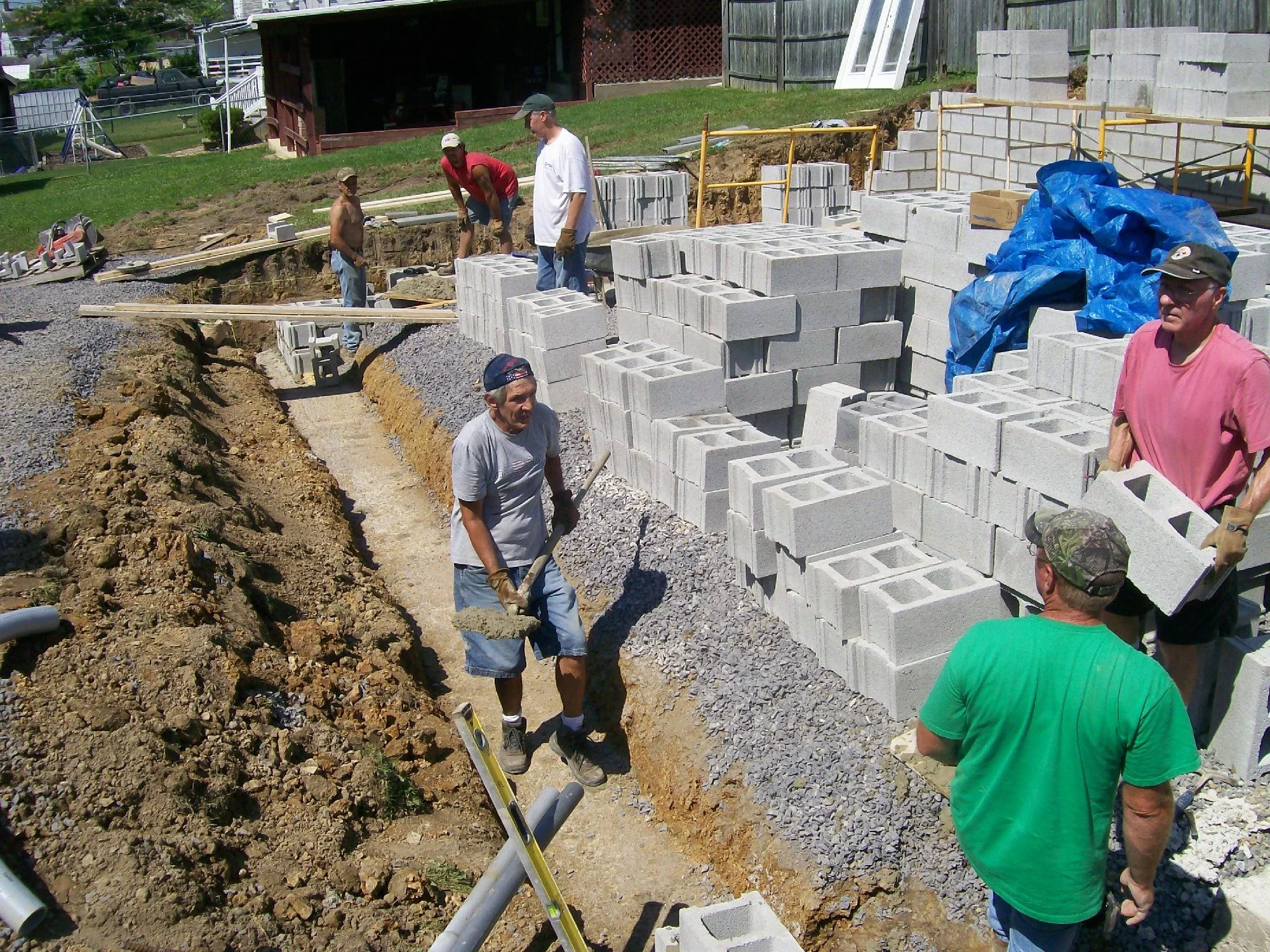Group of construction workers building a concrete block wall outdoors, with some workers handling concrete blocks and others shoveling dirt, in a sunny residential area.