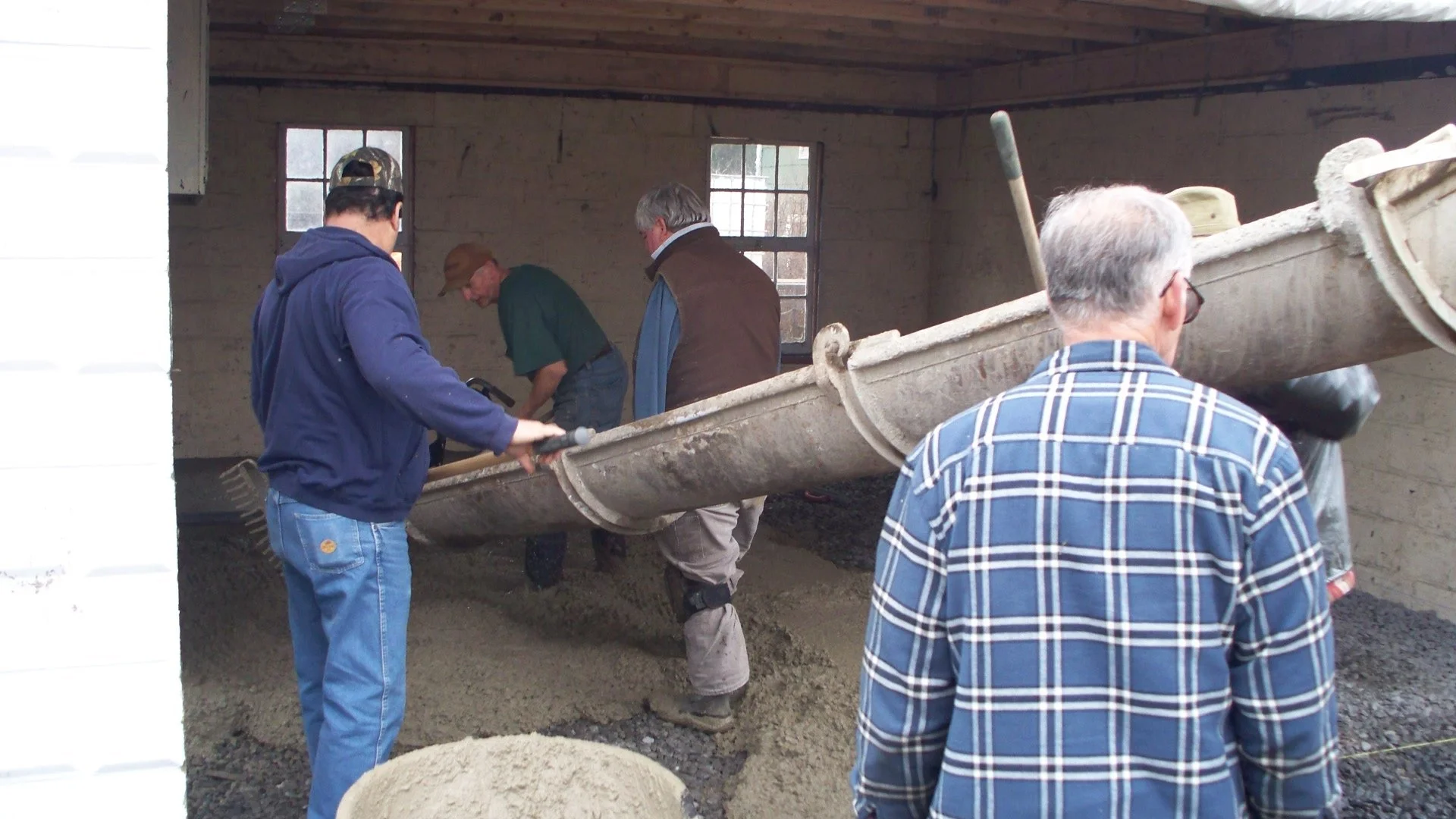 Group of men working together in a barn constructing a small boat, with one man pouring concrete or gravel on the ground.