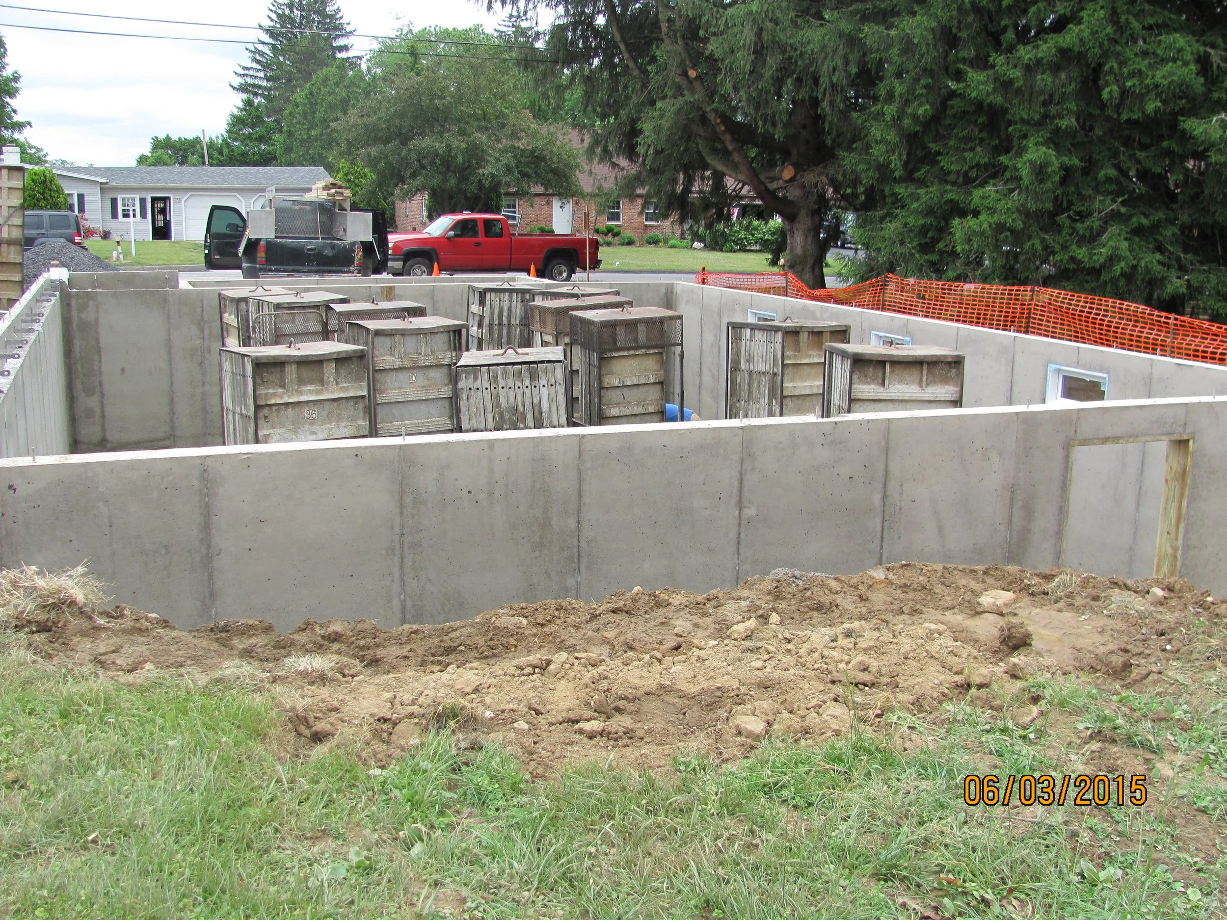 Construction site with a concrete foundation and various construction materials, including wooden crates and a blue plastic container, near residential houses and vehicles. Orange safety fencing surrounds part of the site, with trees and a grassy are