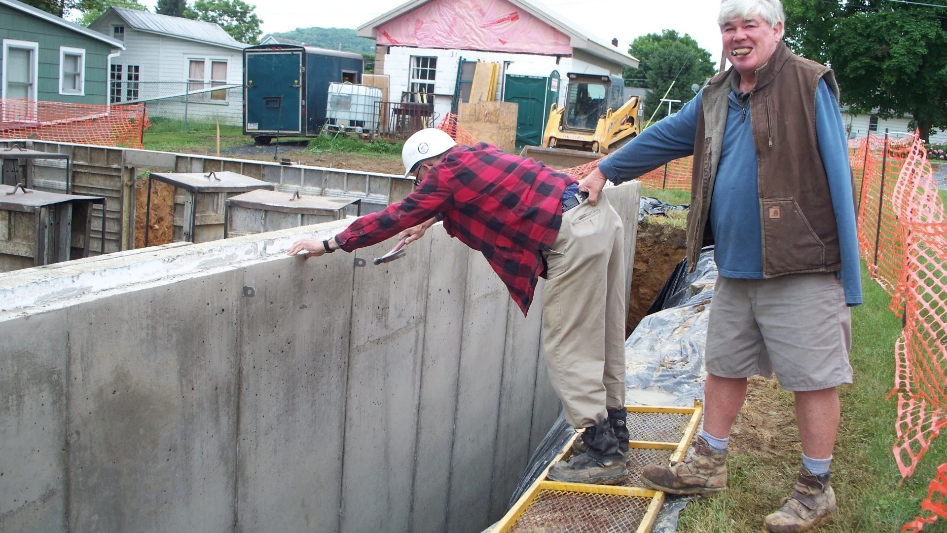 Two men are working on an outdoor construction site next to a concrete foundation wall. One man is leaning over the wall, wearing a white hard hat and red plaid shirt, while the other man is standing nearby, holding the first man by the waist, smilin