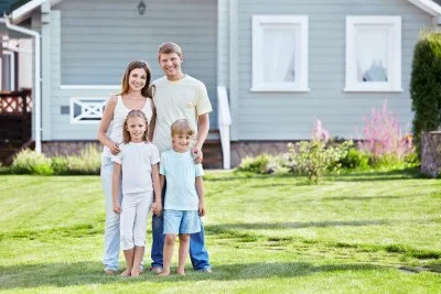A family of four standing outside in front of a blue house with a lawn and flower beds.