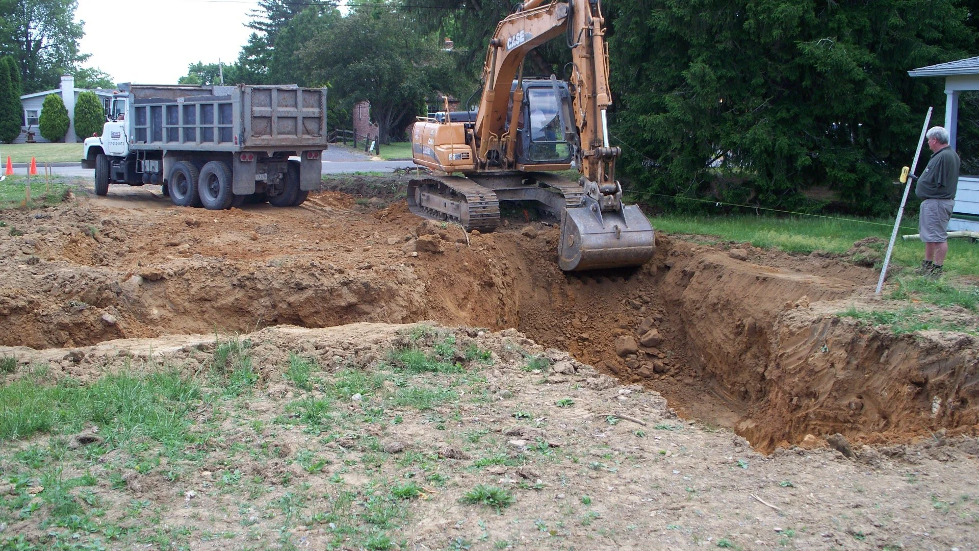 A construction site with a large excavator digging a trench, a dump truck nearby, and a man standing with a leveling tool on the grass to the right.