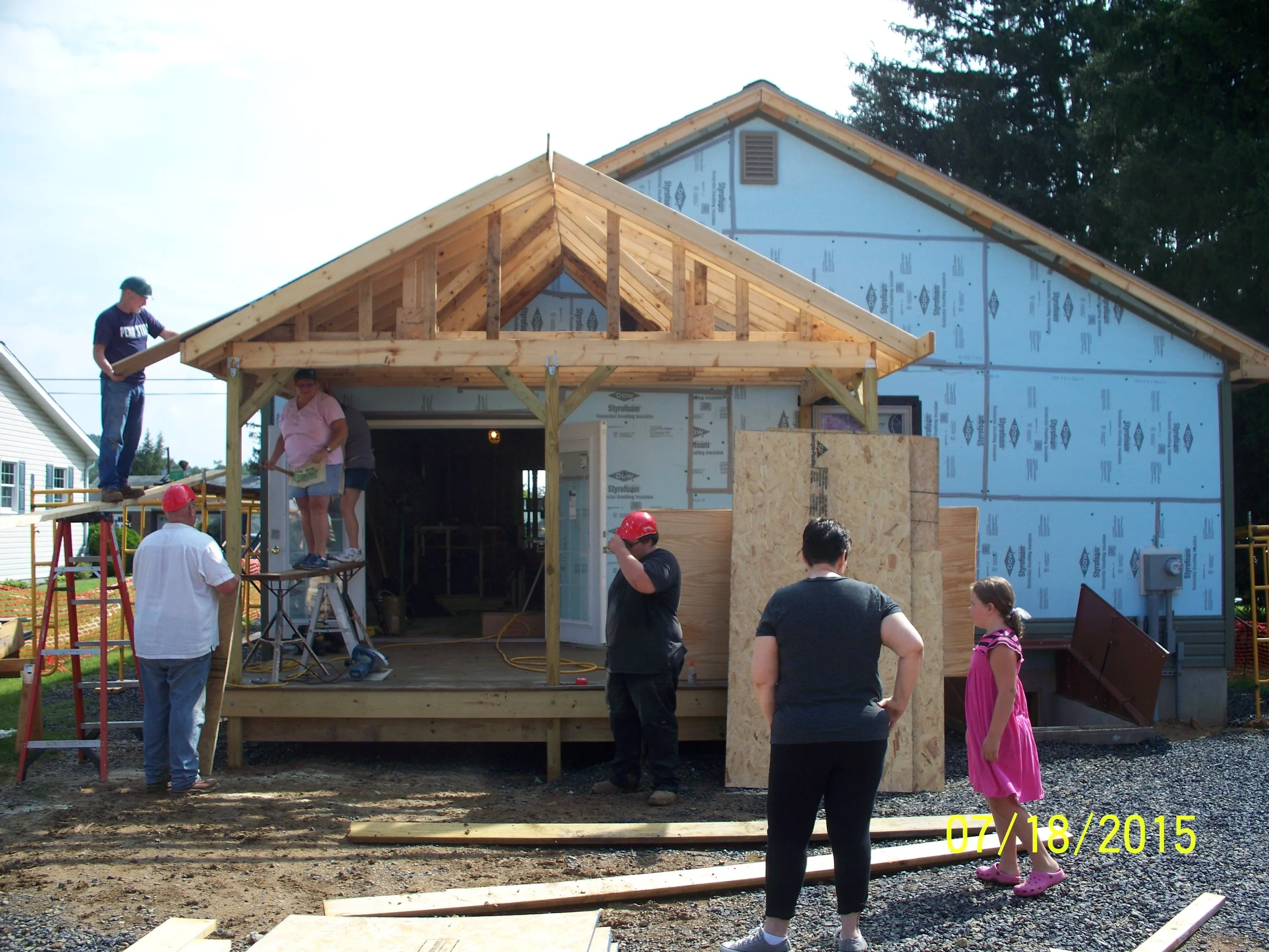 People building a house, with some working on the porch and others observing, under a partly cloudy sky.