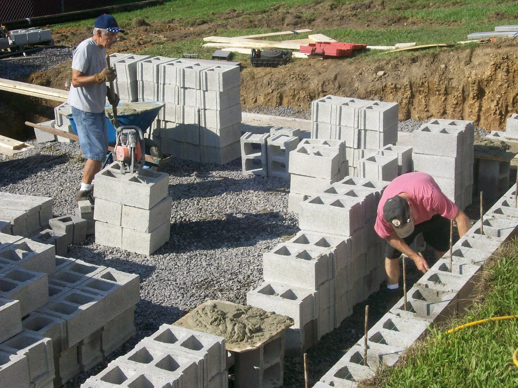 Two men working on constructing a concrete block foundation at a construction site, with stacks of concrete blocks and construction tools around them.