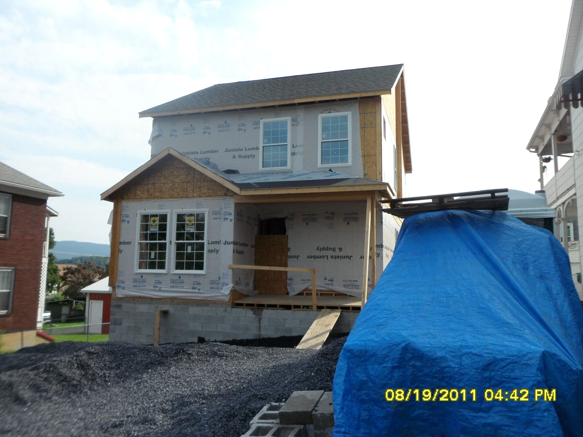 A house under construction with a partially finished second floor, covered in building wrap, and a blue tarp covering a pile of materials in the foreground.