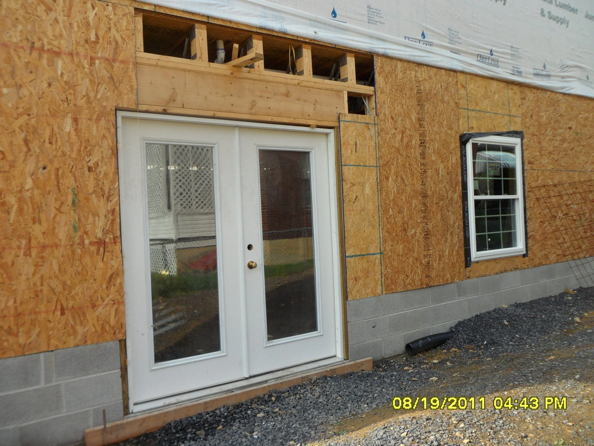 Construction of a building exterior with a white door and window, unfinished wooden framing, and plywood sheathing.